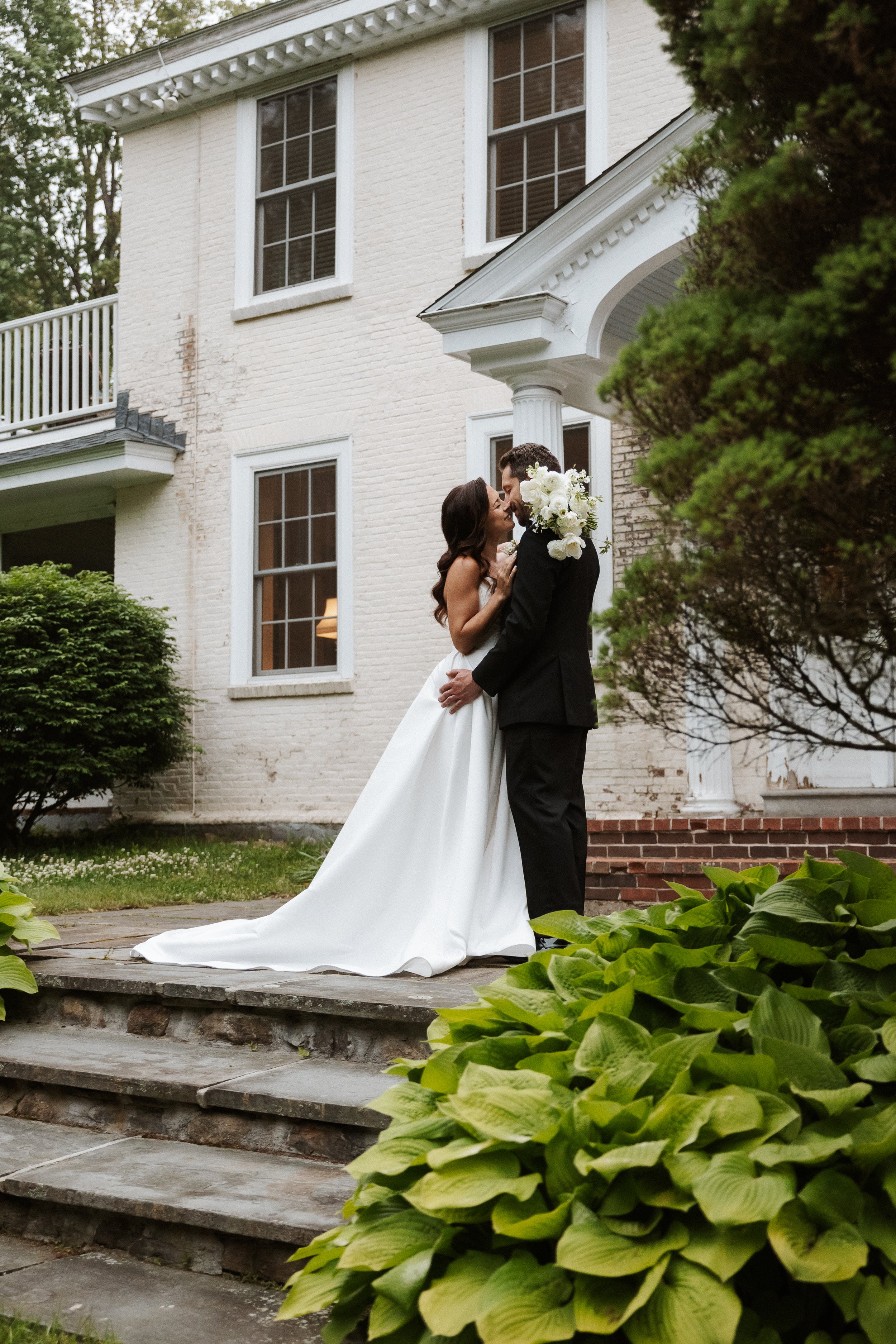 A bride and groom sharing a kiss outside a white brick house. The bride is wearing a white wedding gown, and the groom is dressed in a black suit. The groom is holding a bouquet of white flowers, and they are standing on a stone staircase surrounded