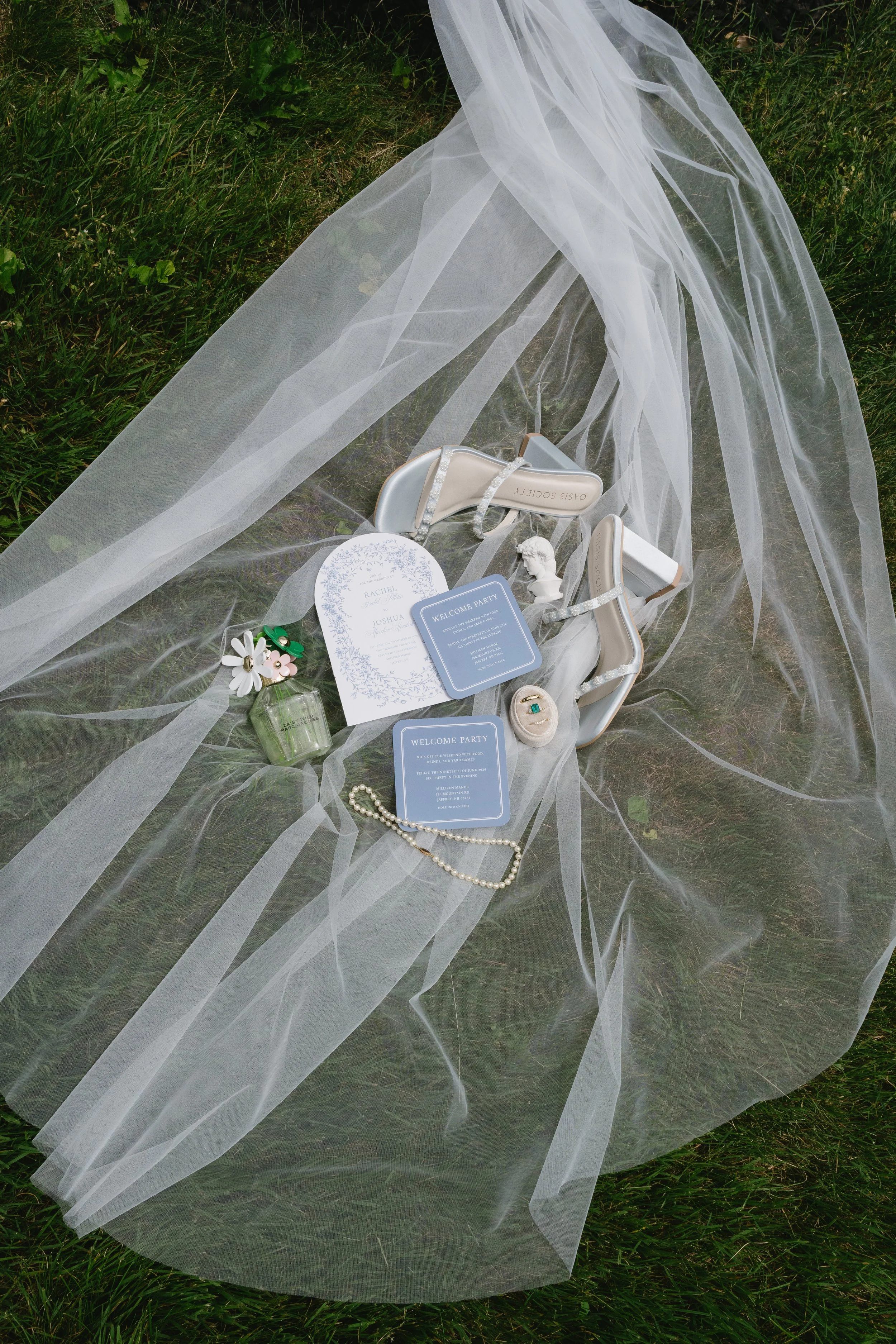 Wedding accessories arranged on sheer fabric on grass, including a pair of cream high-heeled shoes with rhinestones, a small bust sculpture, a pearl necklace, a floral ring, floral hair clips, a bottle of perfume, wedding programs, and a veil.