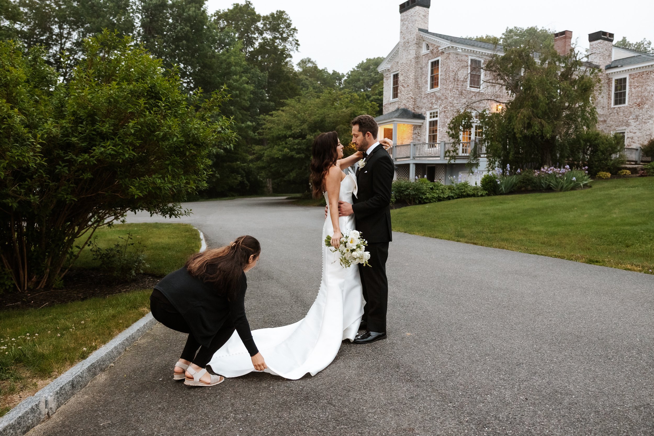 A bride and groom embrace outdoors on their wedding day, with a woman adjusting the bride's long white train, in front of a large brick house surrounded by greenery.