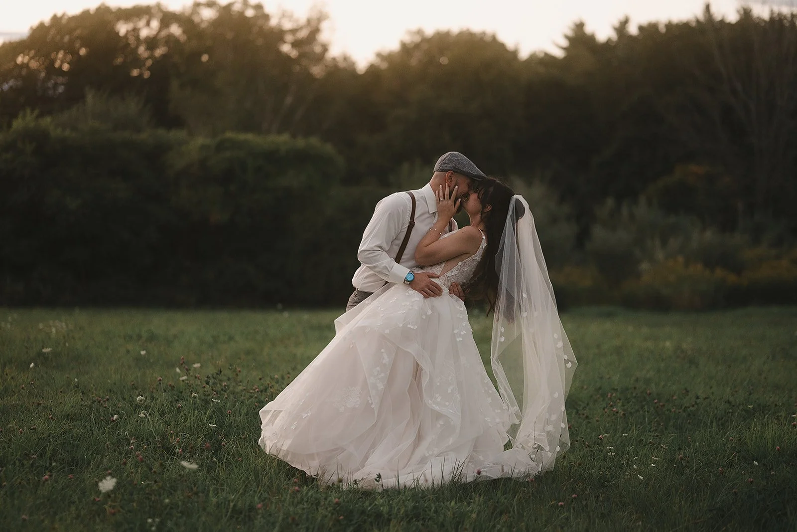 A bride and groom sharing a kiss outdoors during sunset, with green grass and trees in the background, the bride in a white wedding dress and veil, and the groom in a white shirt with suspenders.