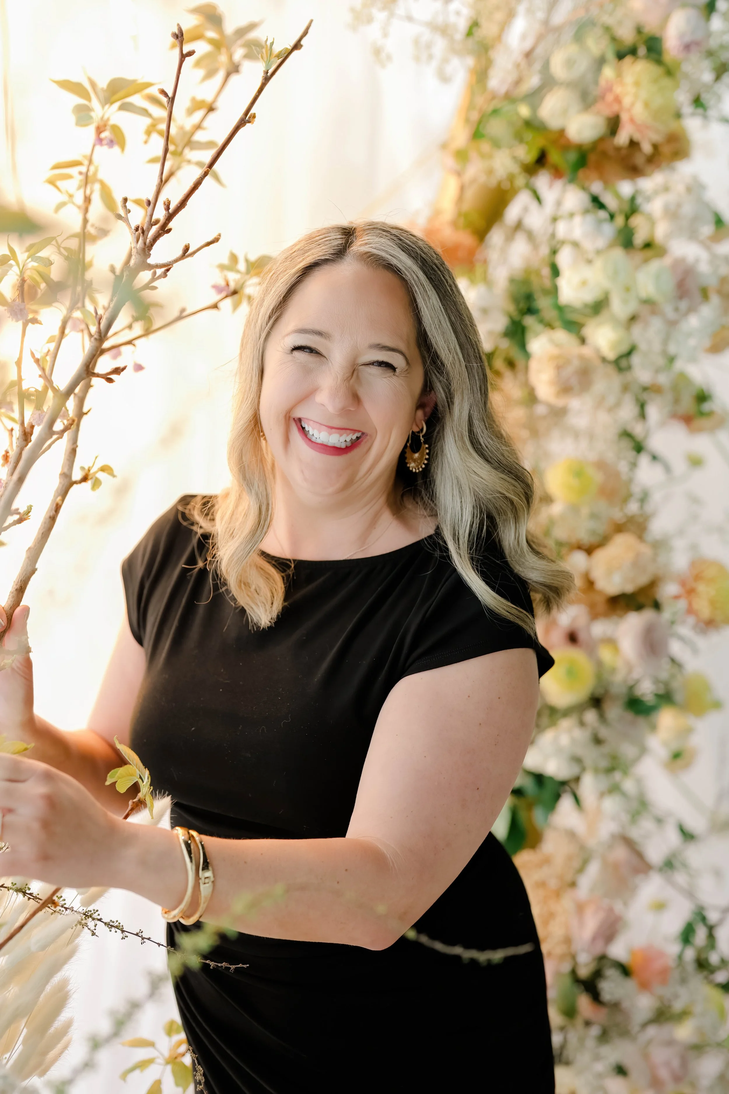 Woman with blonde hair and earrings smiling, surrounded by white and pastel flowers and branches.