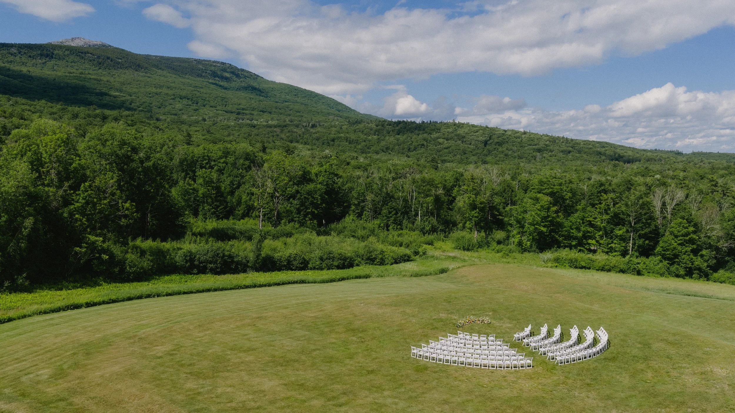 Outdoor setting with rows of white chairs arranged in a semicircle on a well-manicured grassy area, surrounded by lush green trees and hills. The sky is partly cloudy with patches of blue.