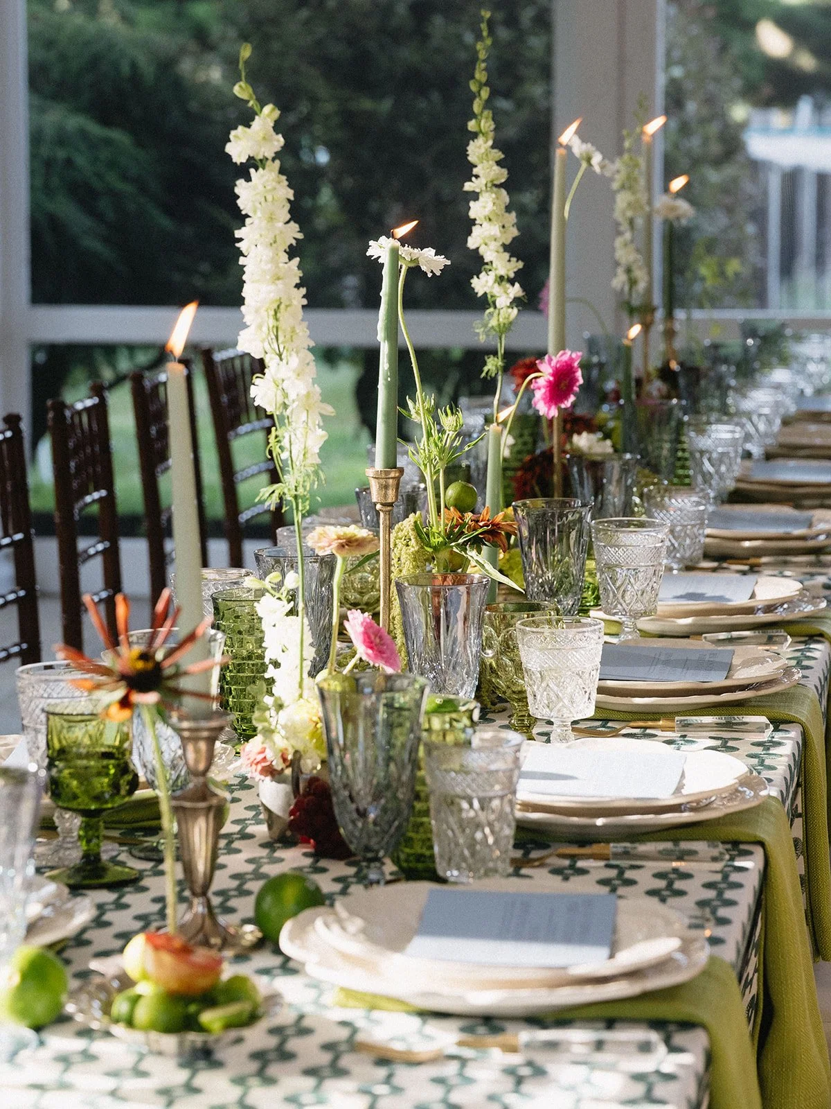 A long dining table set with plates, glasses, and cutlery, decorated with candles and a variety of floral arrangements, with a window showing greenery outside.