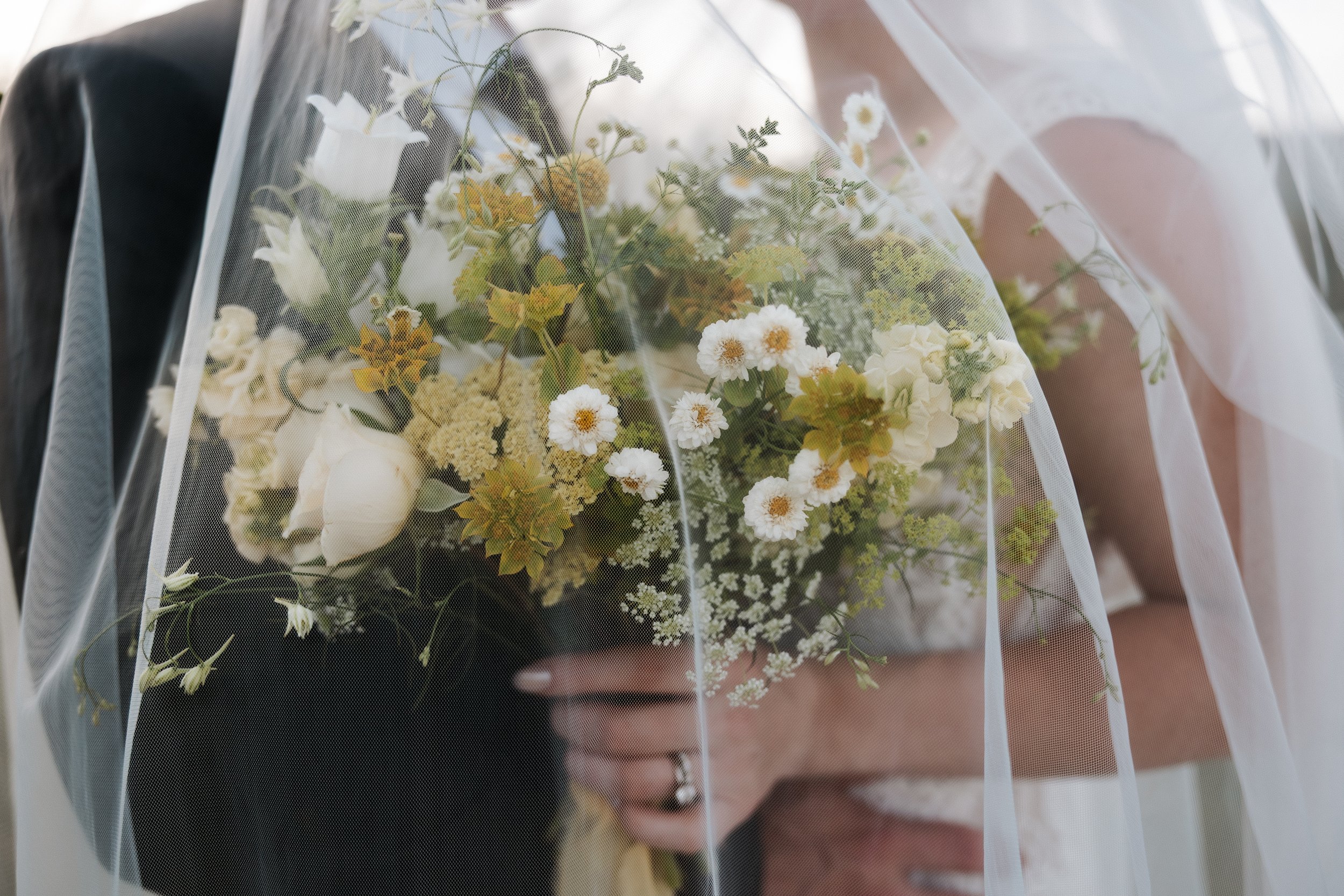 Close-up of a person holding a wedding bouquet with white flowers and greenery, partially covered by a sheer veil.