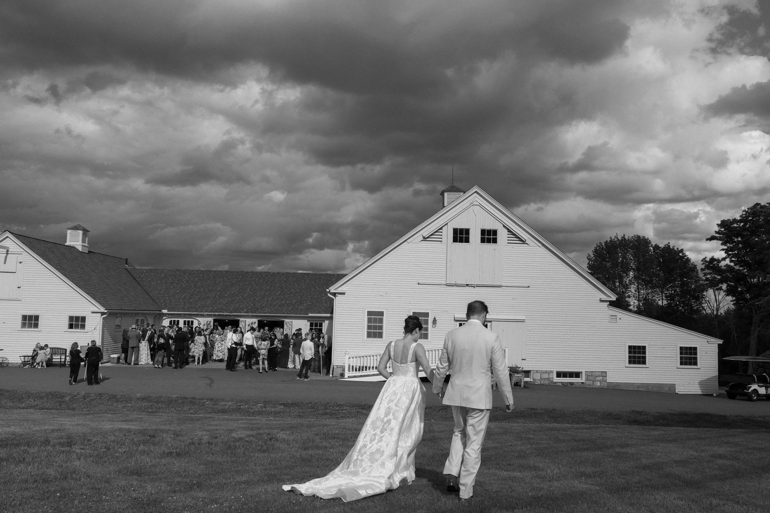 A bride and groom walking on grass outside a large white barn with dark clouds above at a wedding reception.