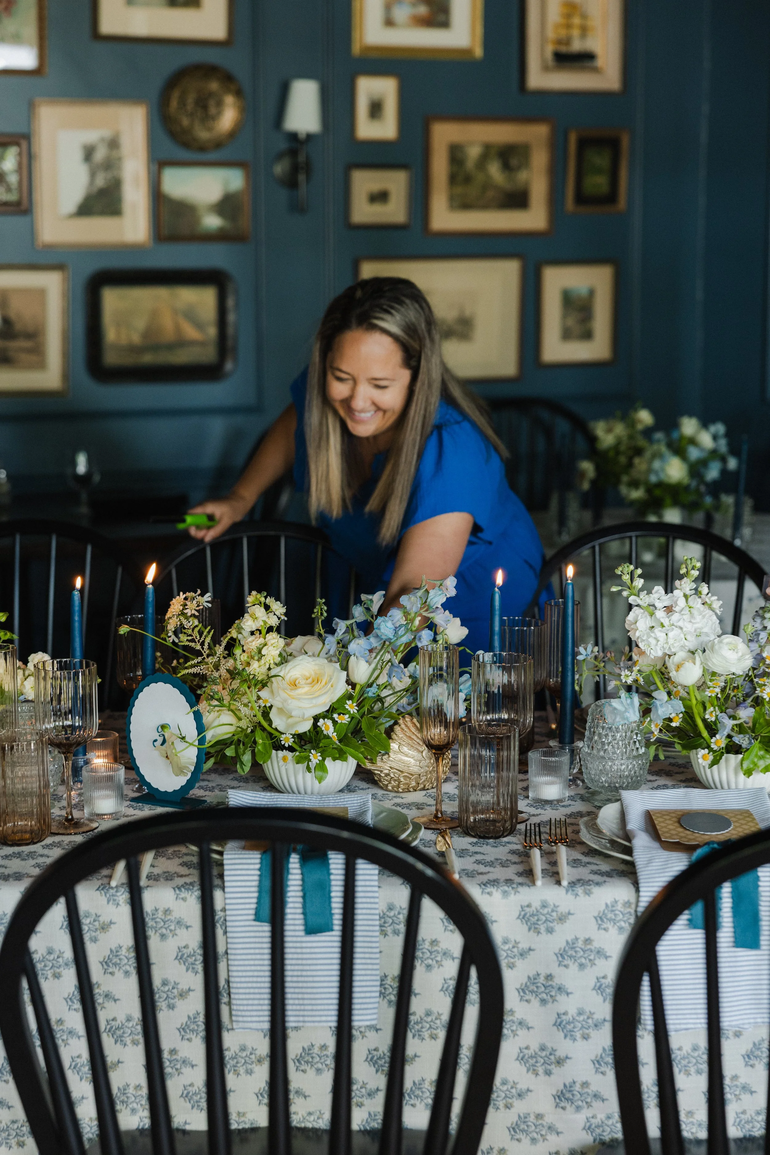 A woman with long brown hair in a blue dress is smiling and leaning over a table decorated with white and blue floral arrangements, candles, and tableware. The setting is a dining room with teal walls, decorated with numerous picture frames, and modern black chairs.