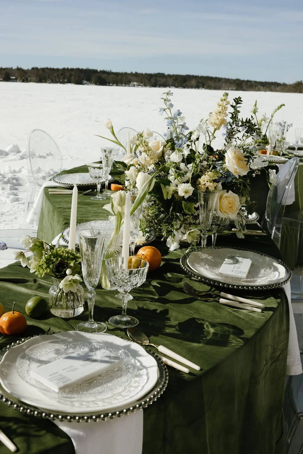 A table set for an outdoor winter event with green tablecloth, white and green floral centerpiece, glassware, silverware, candles, and orange fruits, overlooking a snowy landscape.