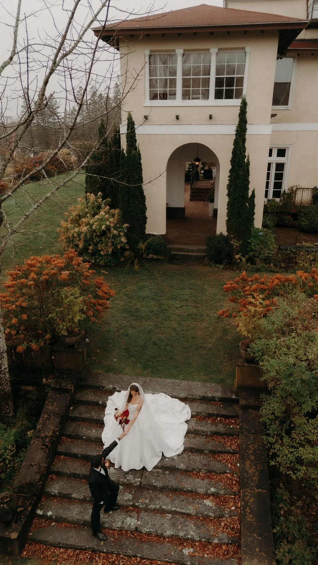A bride in a white wedding gown descending outdoor stairs and holding a bouquet while a groom in a black suit lifts her hand.