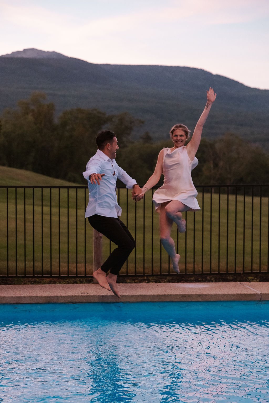 A couple jumping into a swimming pool holding hands, with a mountain and trees in the background during sunset.