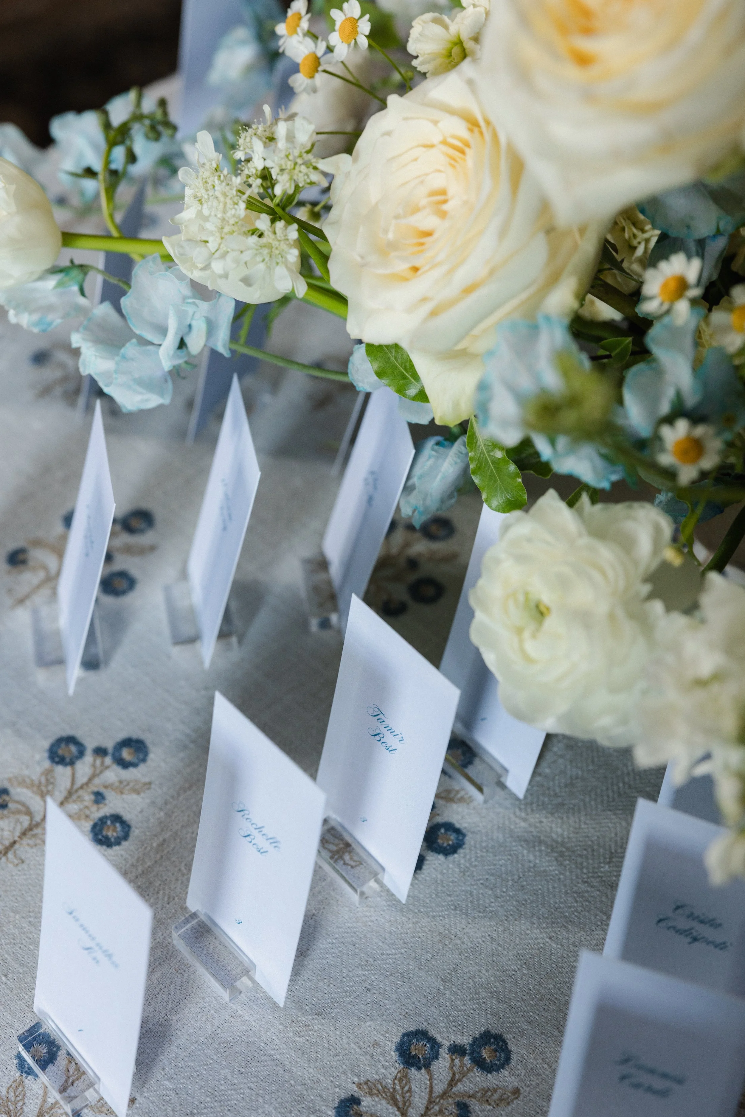 A close-up of a floral arrangement with white roses, daisies, and other small flowers placed on a table with name tags in front of each flower.