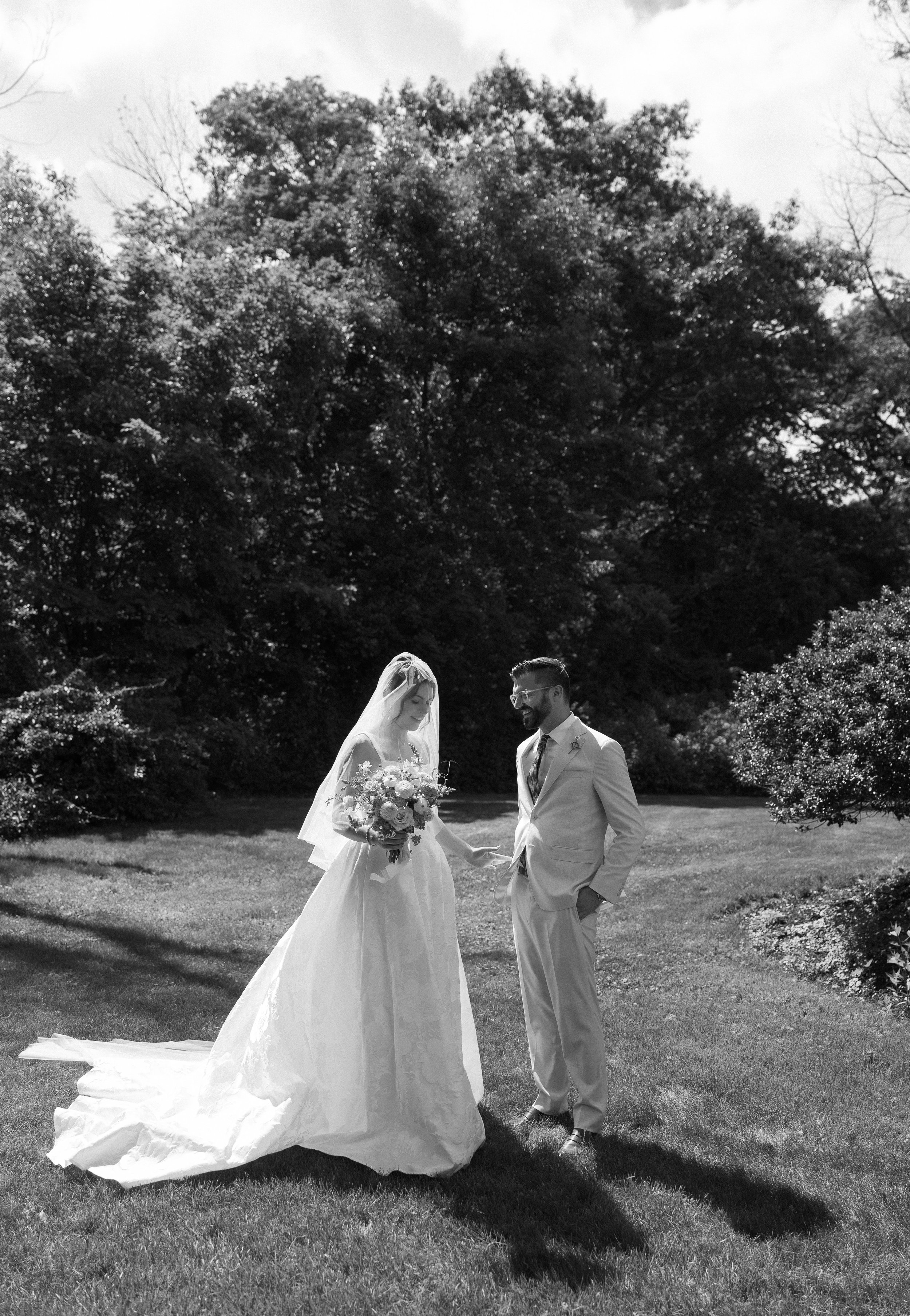 Black and white photograph of a bride and groom standing on a lawn with large trees in the background. The bride wears a wedding gown with a long train and veil, holding a bouquet of flowers. The groom is dressed in a light-colored suit and glasses, smiling at the bride.