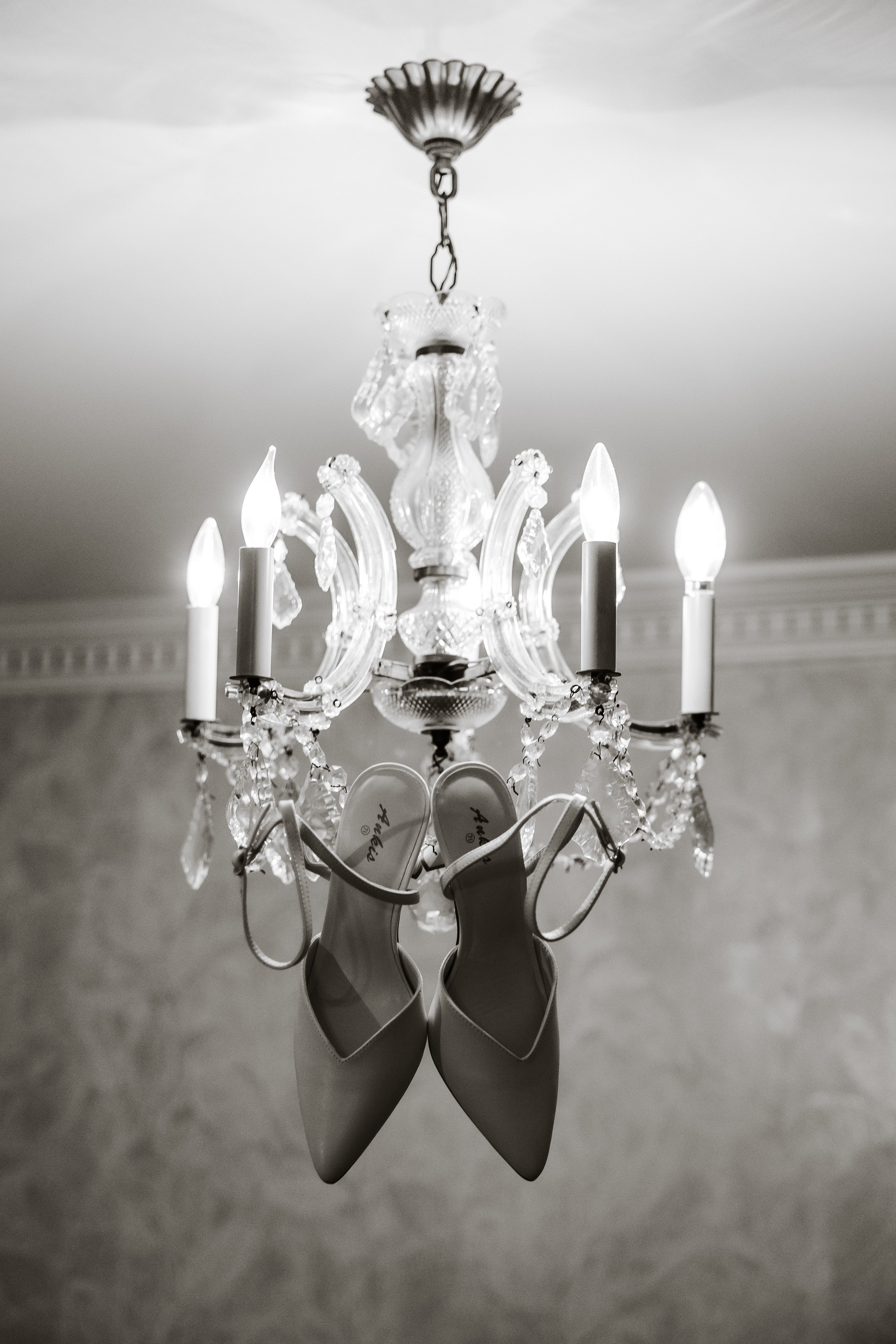 A black and white image of a chandelier hanging from the ceiling with a pair of white high-heeled shoes hanging from it.