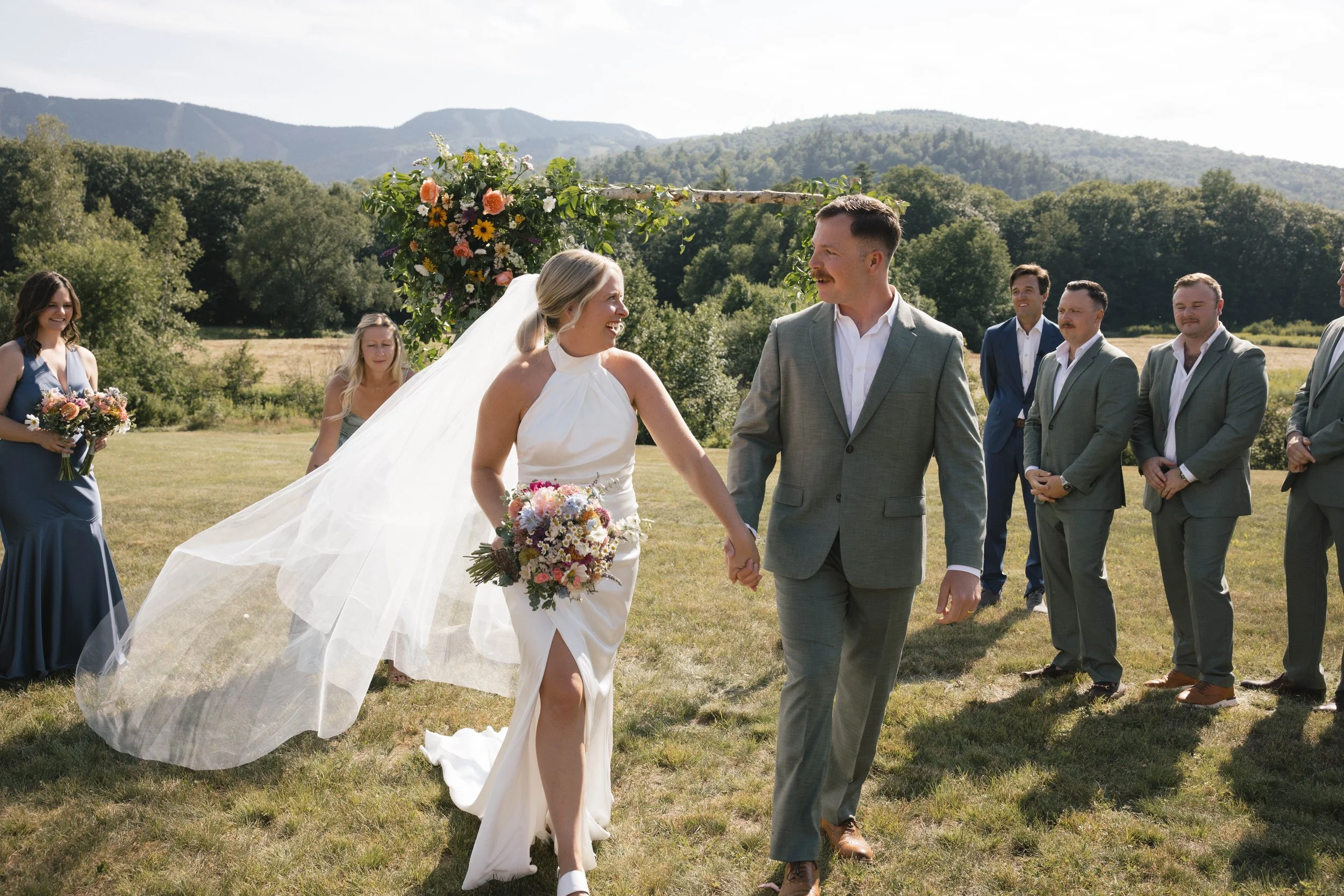 A wedding ceremony outdoors with a bride and groom holding hands and smiling at each other, surrounded by friends and family, with mountains and trees in the background.