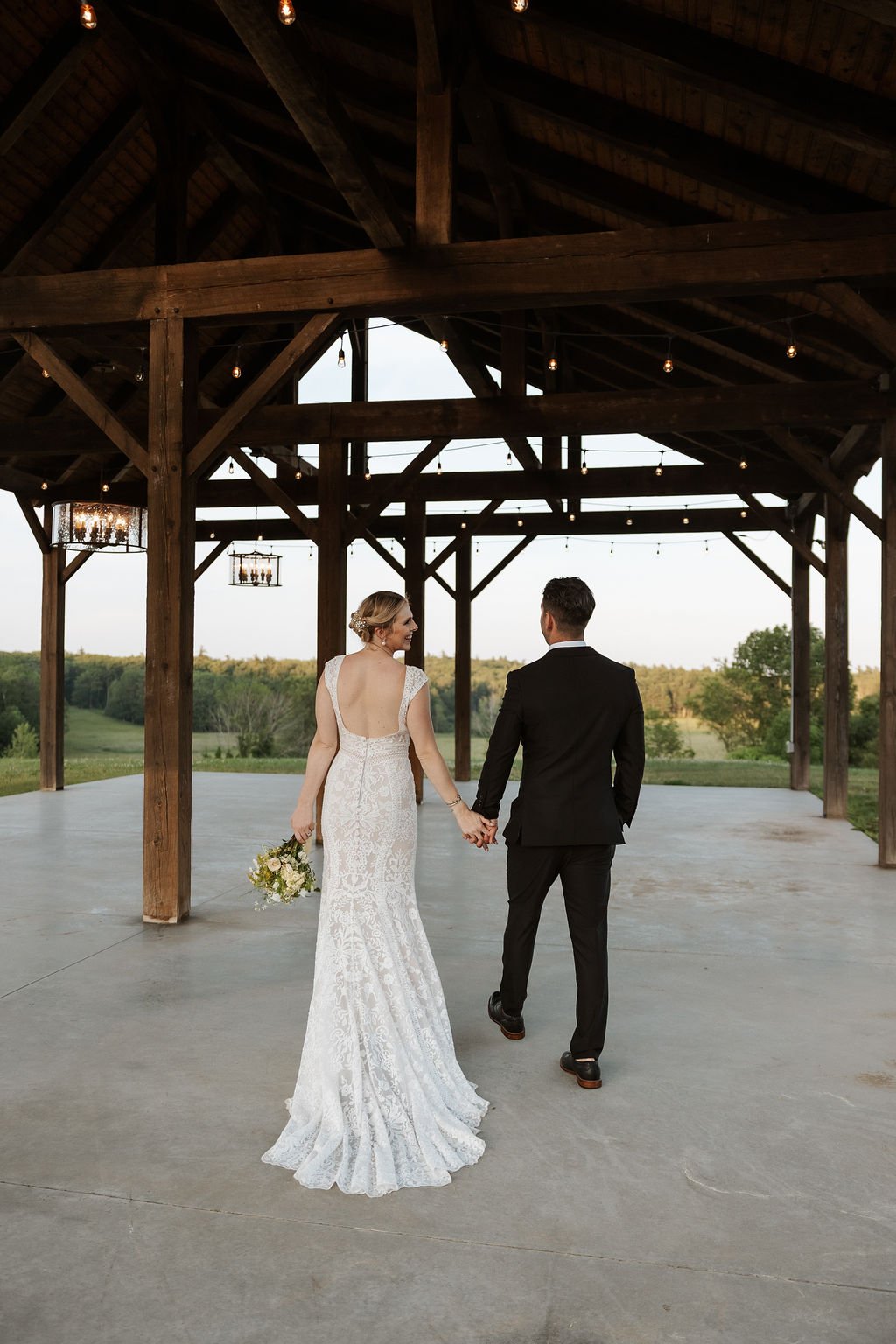 Bride and groom holding hands, walking away in an open wooden pavilion with string lights, during sunset, in a rural landscape.