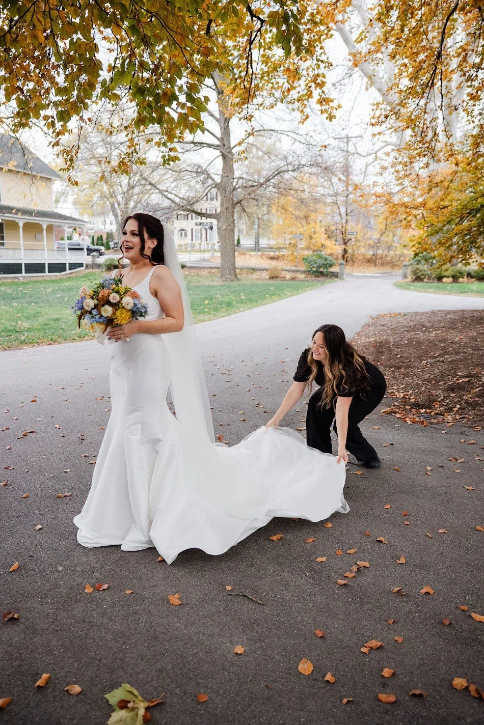 A bride in a white wedding dress holding a bouquet of flowers, standing on a paved path in a park during autumn. A woman in black, likely a helper or stylist, adjusts the train of the dress. The background features trees with orange and yellow leaves, a house, and a street.