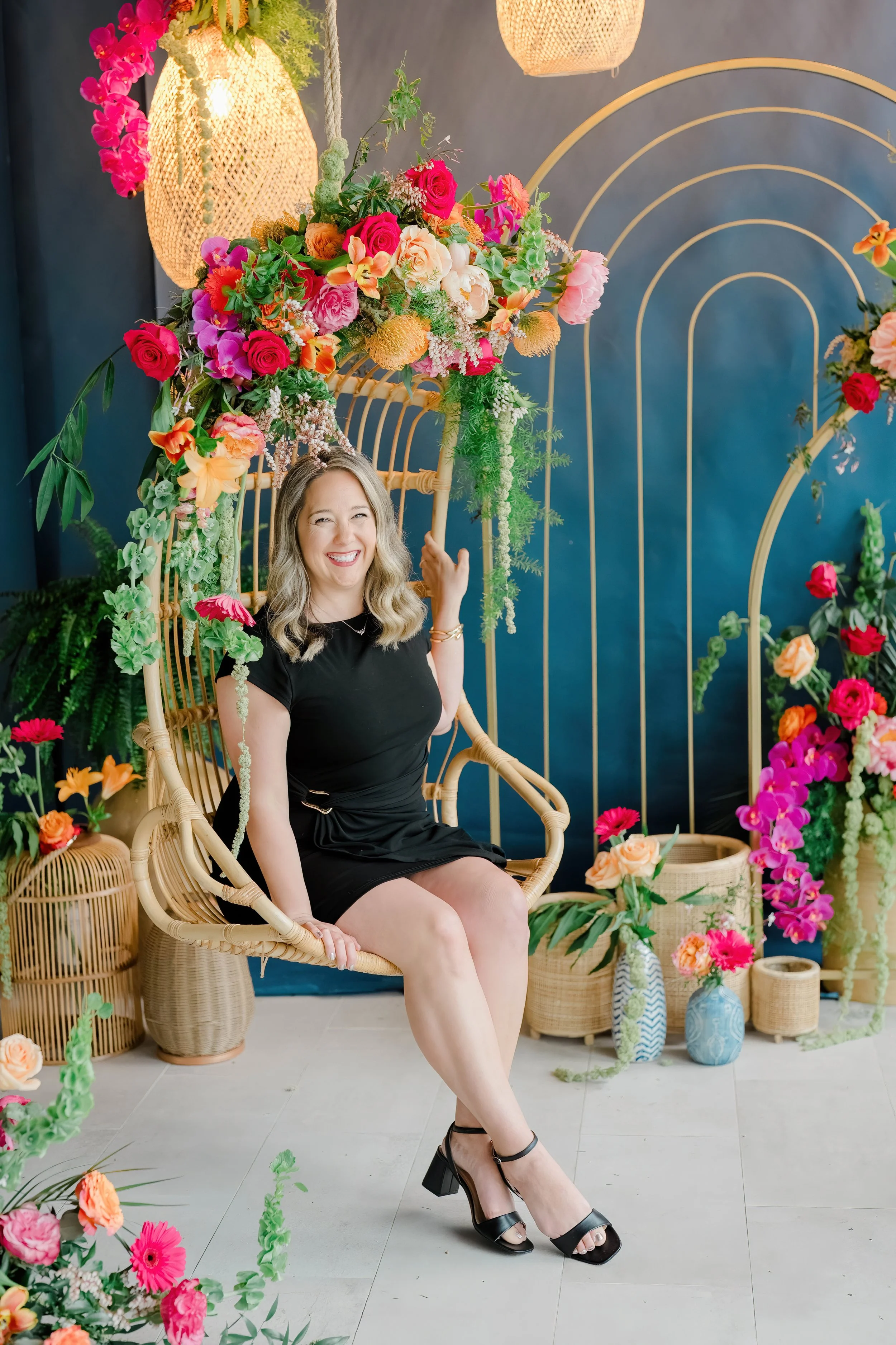 A woman sitting in a rattan chair surrounded by colorful flowers and decorative vases, smiling and wearing a black dress and black high-heeled shoes.