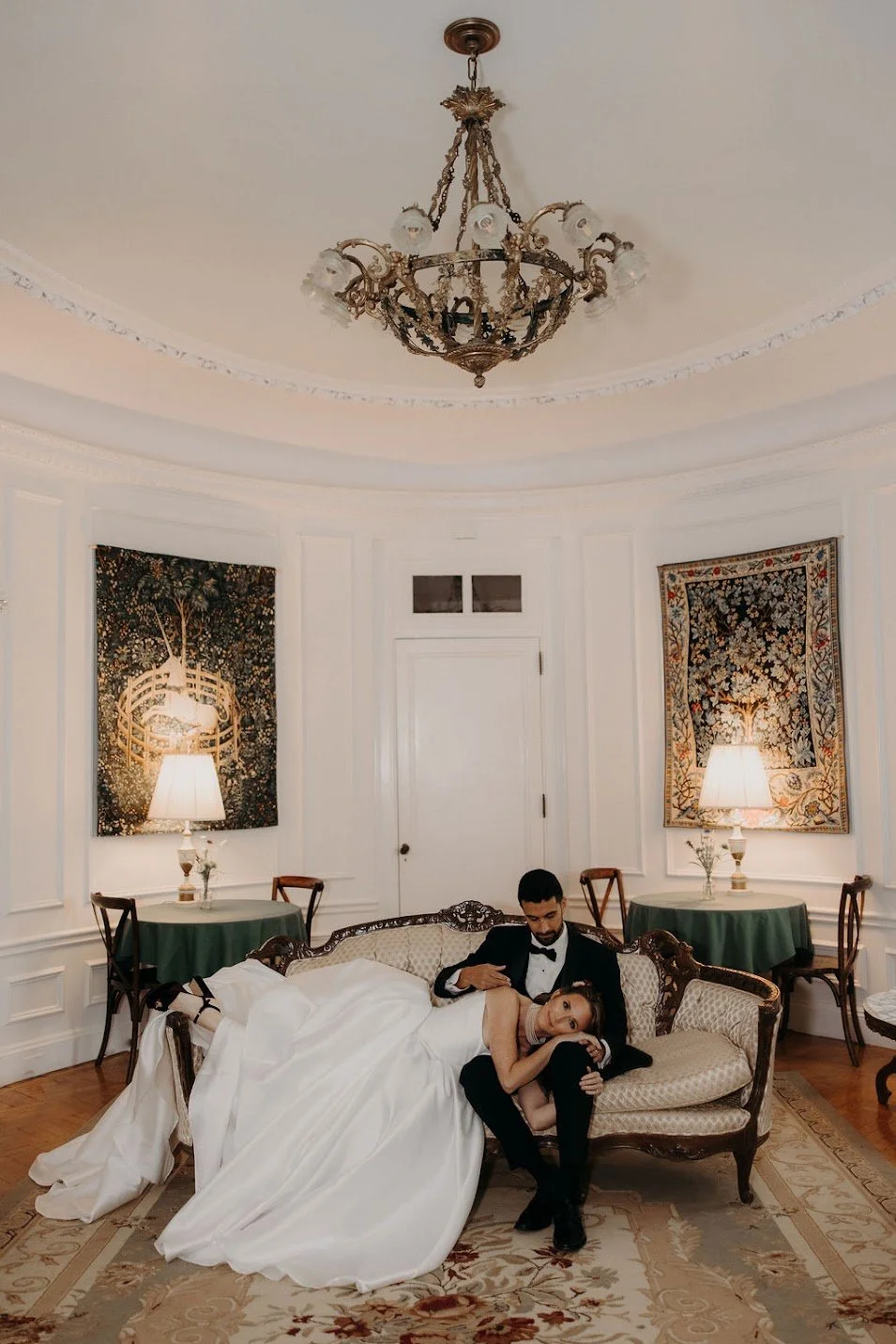 A bride and groom sitting on a vintage sofa in an elegant, classic room with white paneled walls, green-draped tables, lamps, and tapestry wall hangings. The bride lies on the groom's lap, and they are dressed in formal wedding attire.