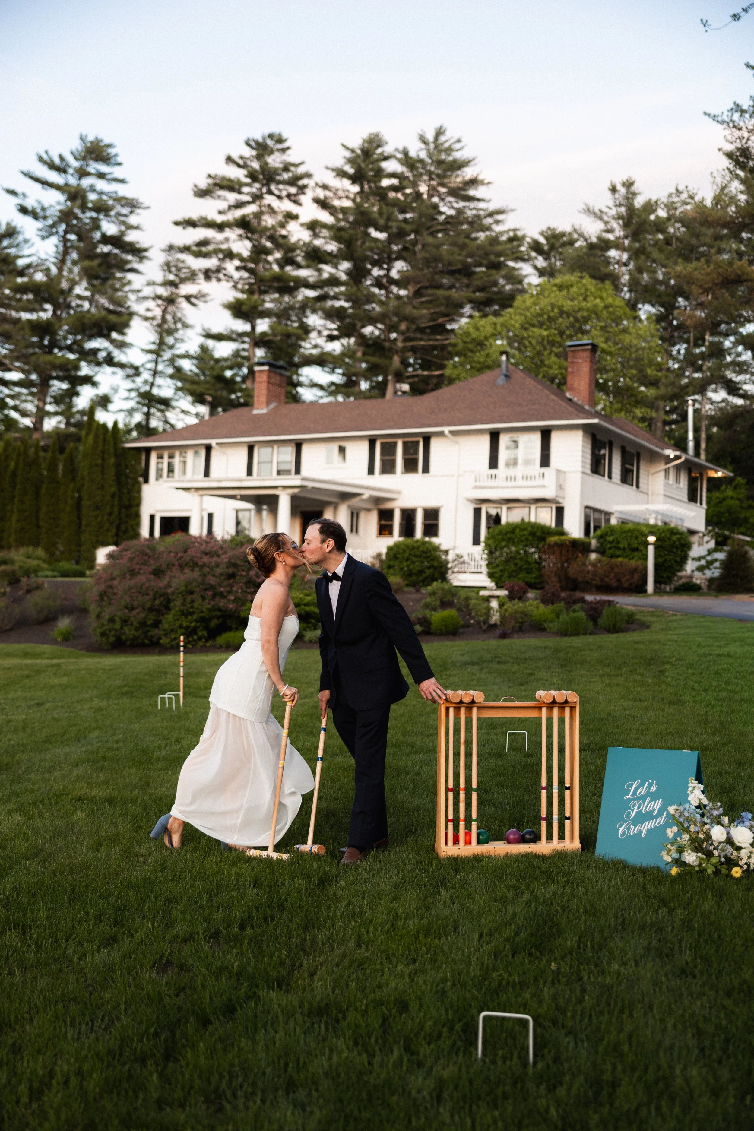 A bride and groom playing croquet in front of a large white house surrounded by lush greenery and trees, during sunset.