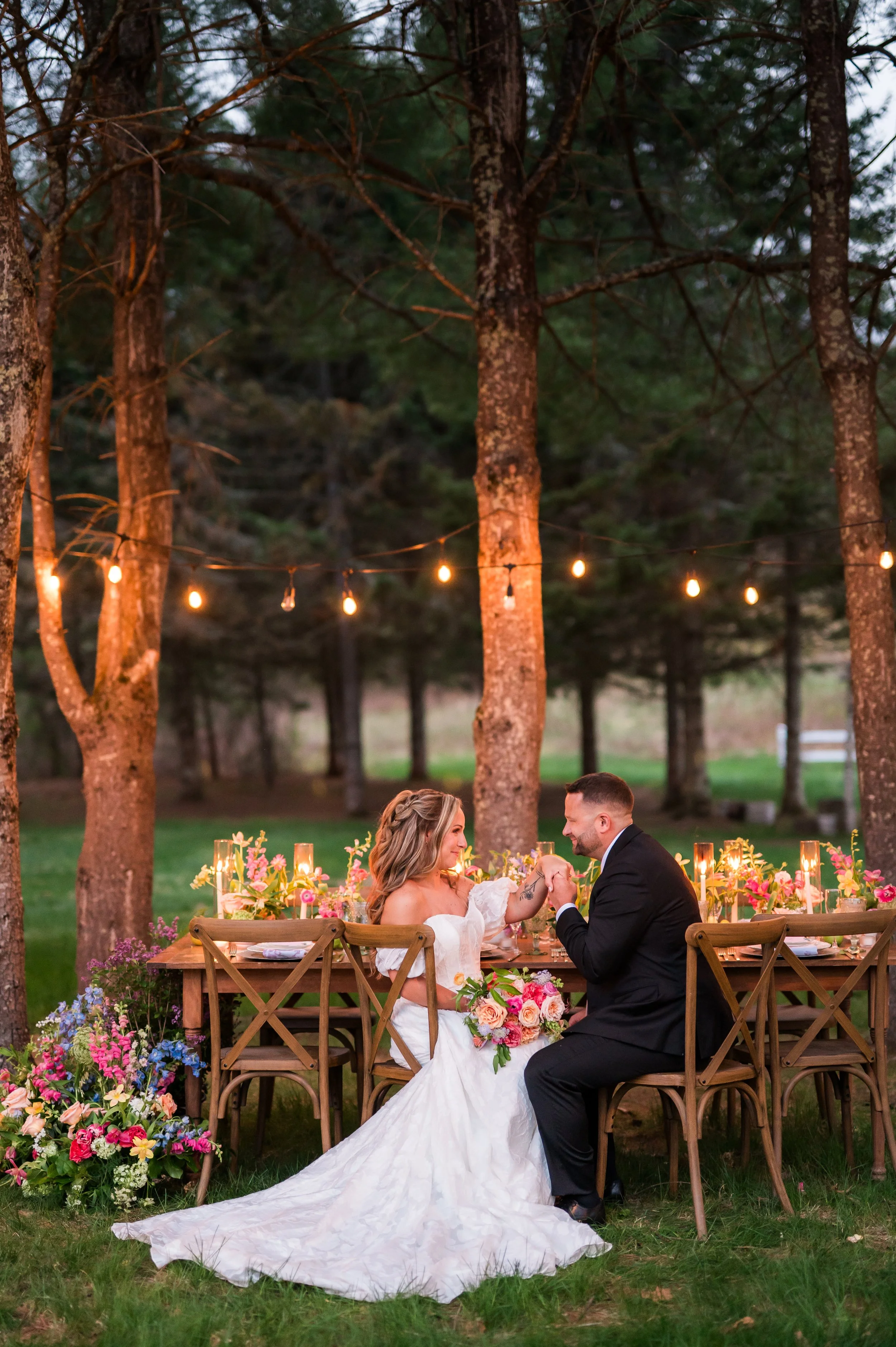 A bride and groom sitting at a wooden outdoor wedding table, holding hands, with a decorated floral centerpiece and fairy lights hanging among tall trees at dusk.