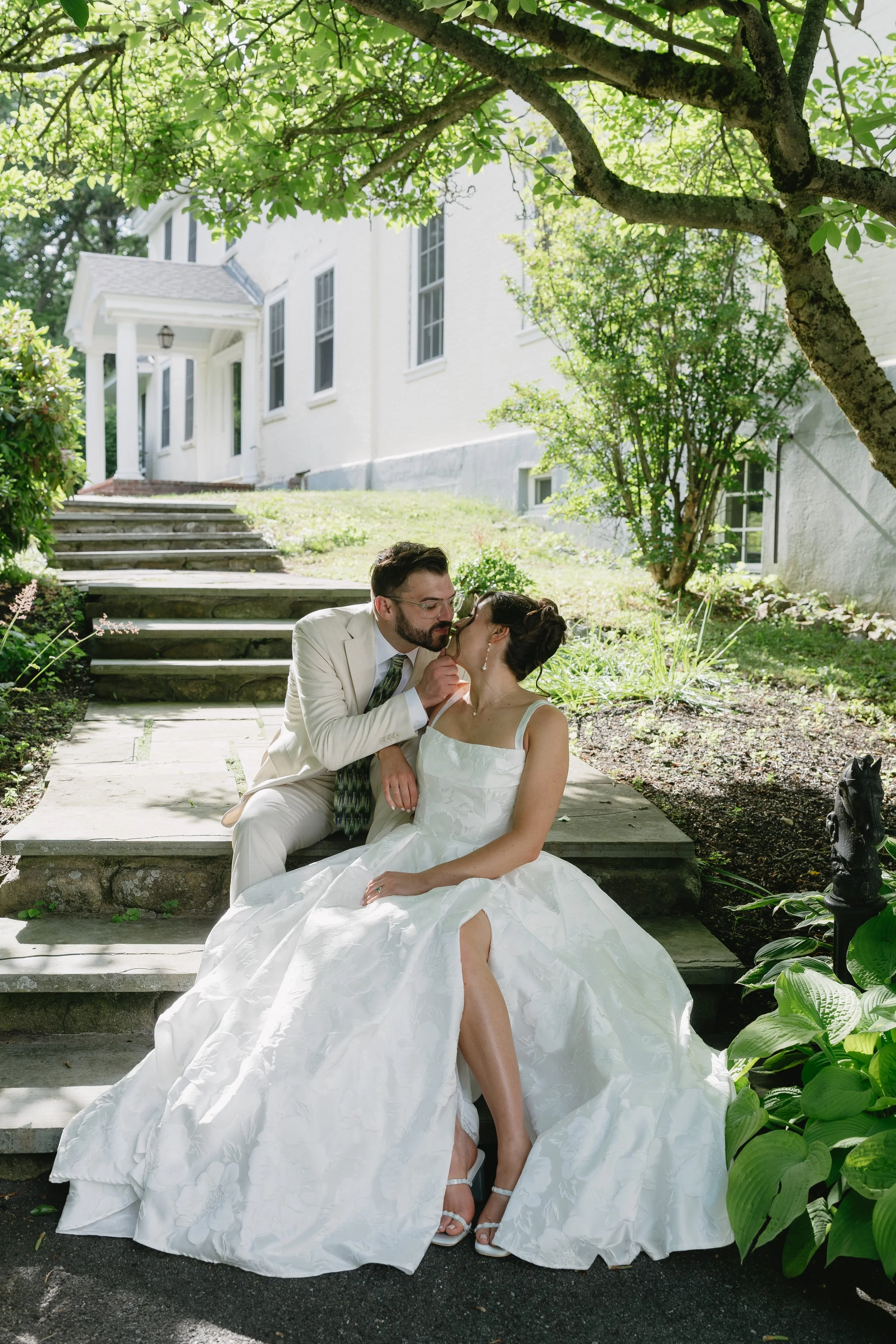 A bride and groom sitting on stone steps outdoors, surrounded by greenery and trees, with a white house in the background. The bride is wearing a white wedding dress, and the groom is in a light-colored suit.