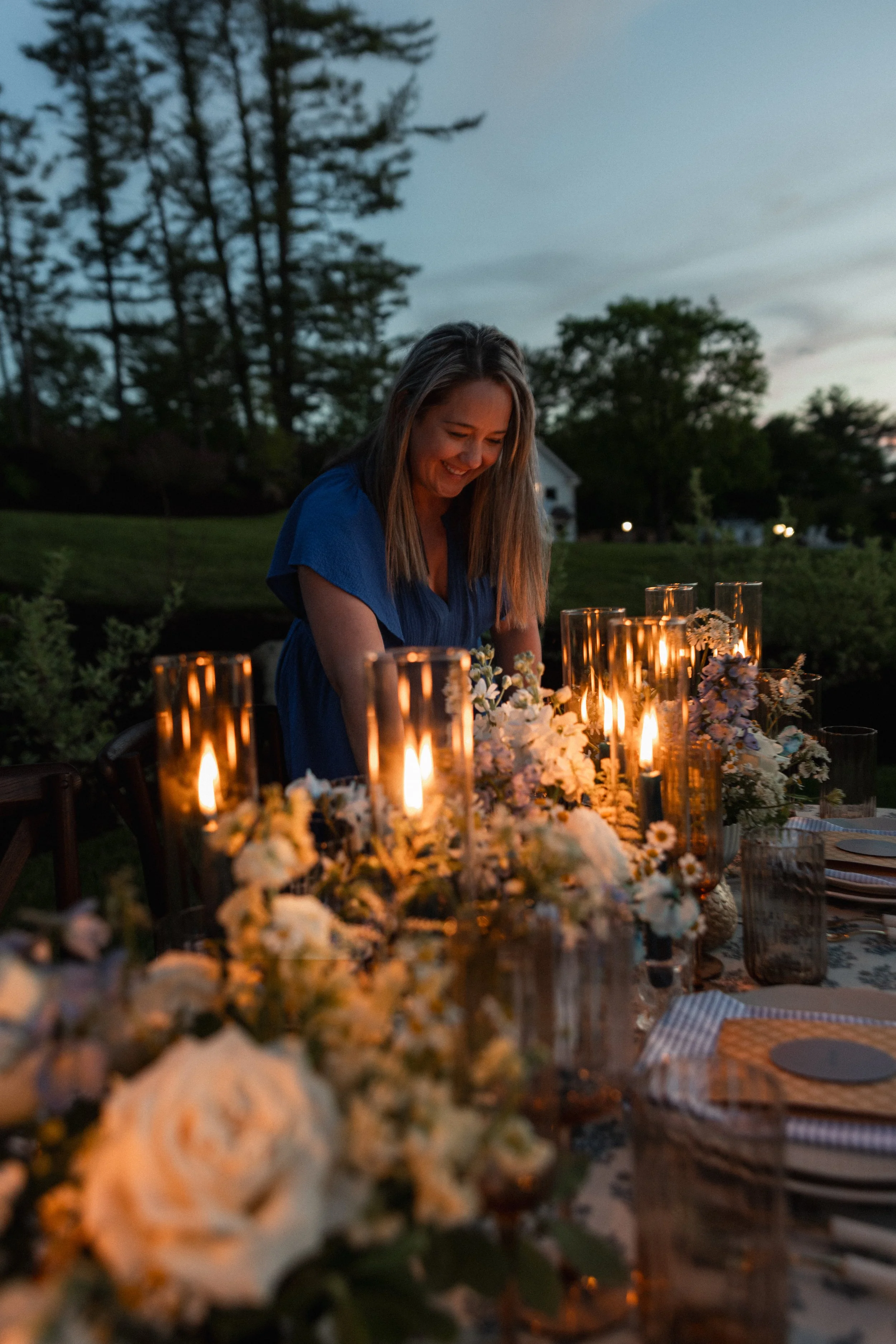A woman in a blue dress smiling at an outdoor dinner table illuminated by candles during dusk, with trees and a house in the background.