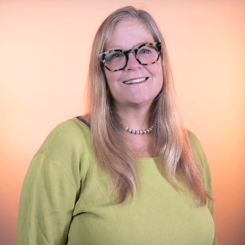 A white woman with long blond hair wearing glasses and a long sleeve green shirt against a light orange background