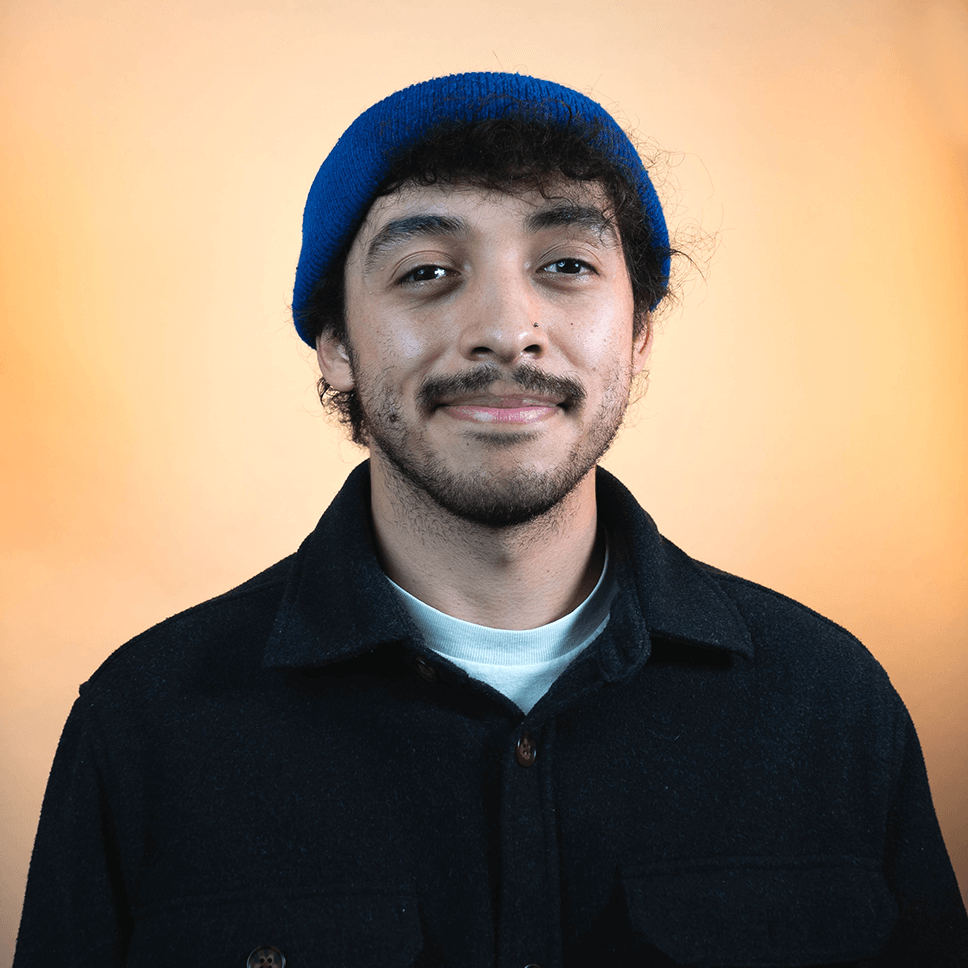 A Latino man with short brown hair and a beard is wearing a blue hat and dark blue button up shirt smiling into the camera