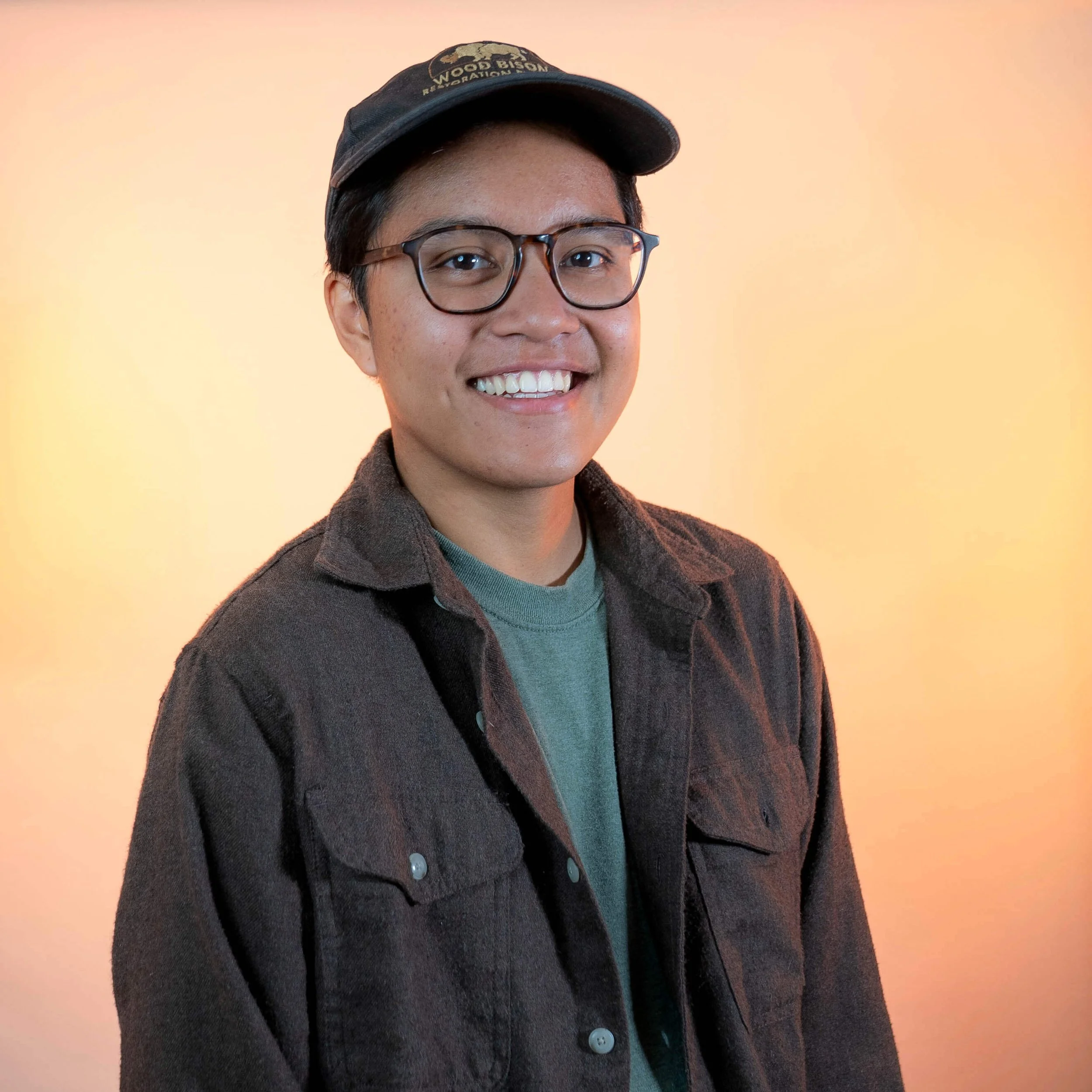 A nonbinary person with short dark hair is smiling and wearing a baseball cap and glasses and a dark grey button down shirt with a green t-shirt underneath and standing against a light orange background