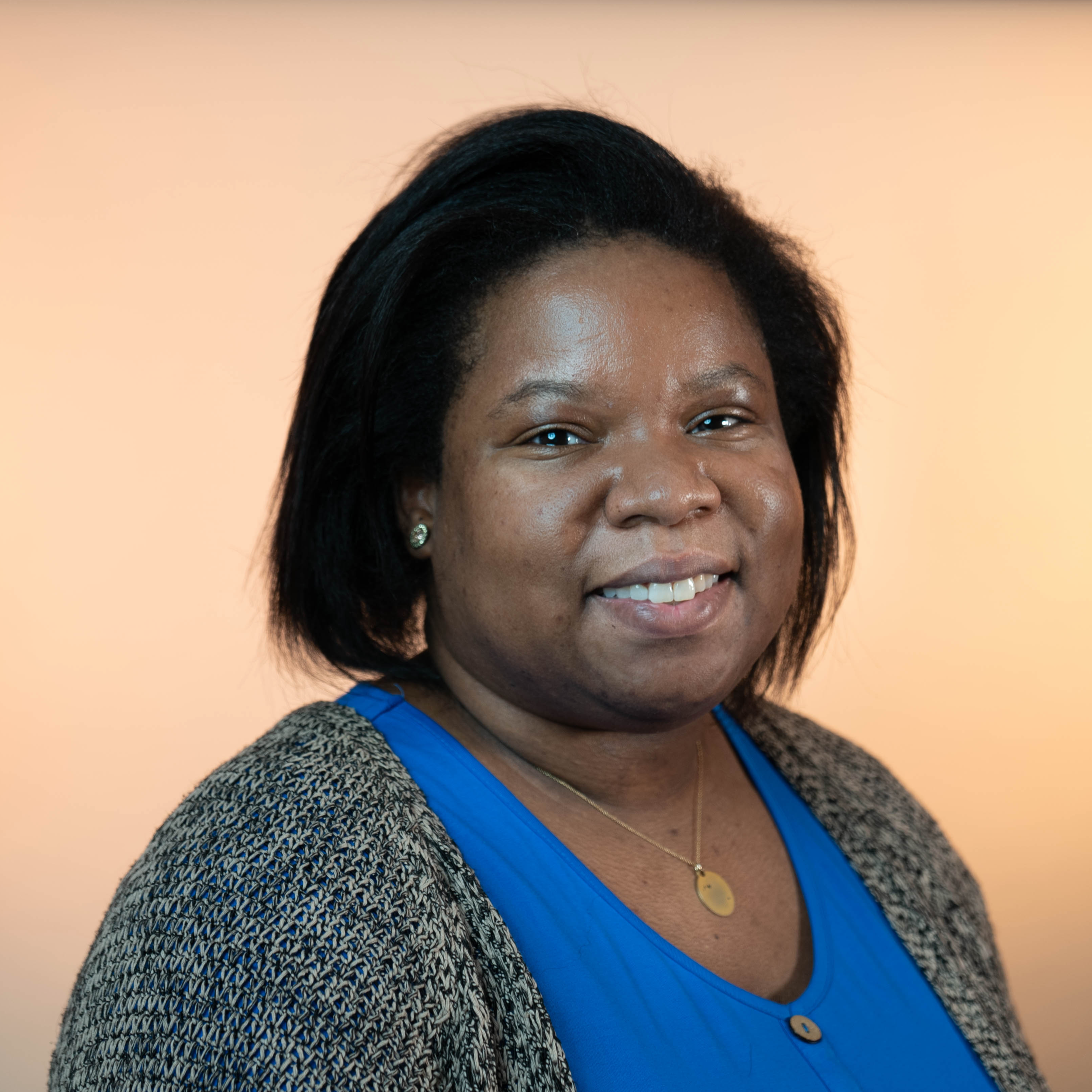 A Black woman with shoulder length dark brown hair is smiling and wearing a grey cardigan over a bright blue shirt with buttons standing against a light orange background