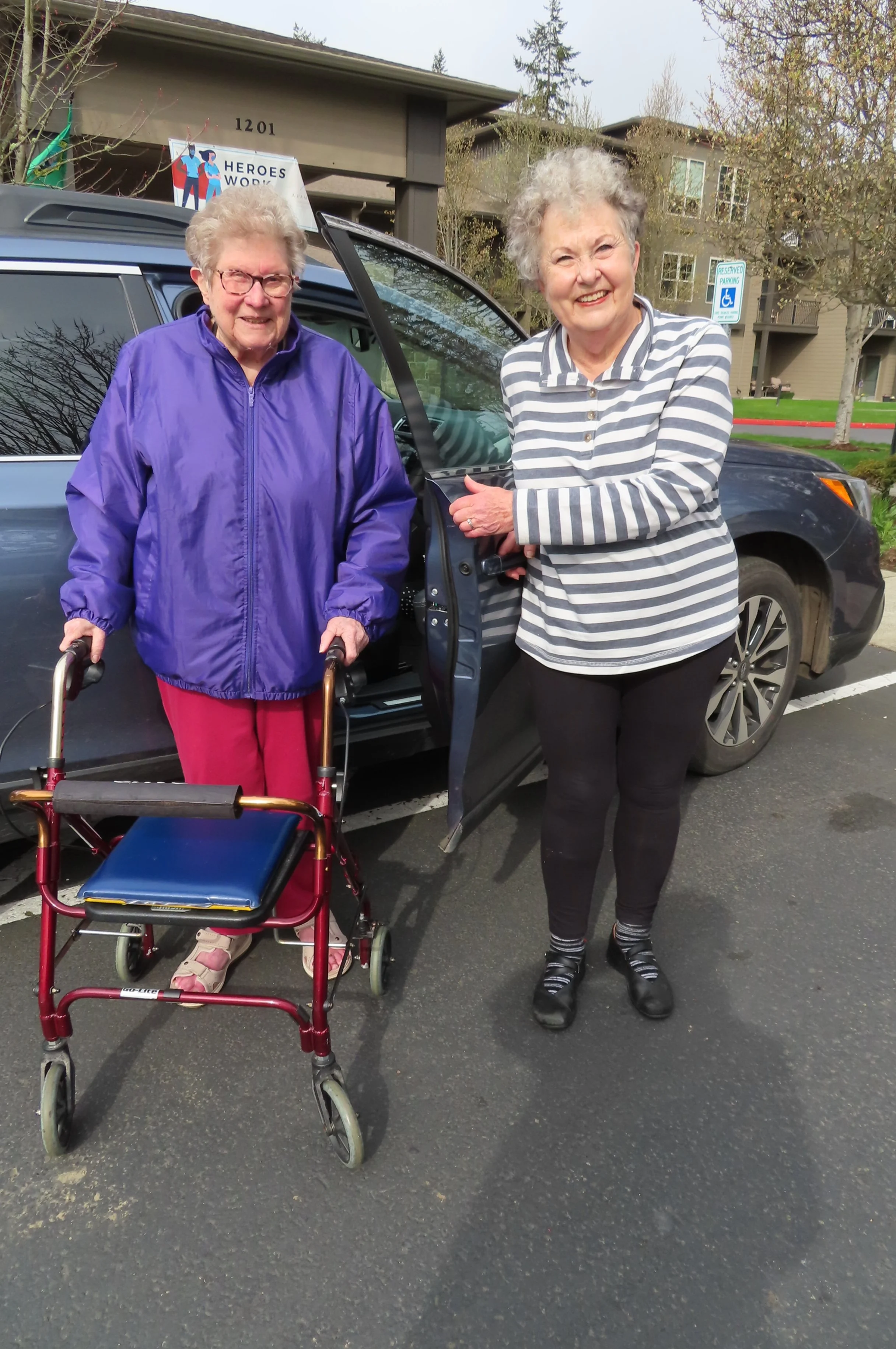 Photo of Volunteer Driver Margie, and a client with a four wheeled walker getting out of a car and standing. They are smiling, and Margie is holding the door.