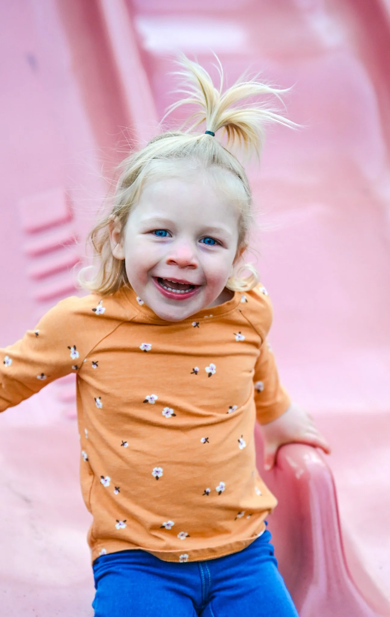 Child with blonde hair sitting on a pink slide, wearing an orange floral shirt and blue pants, smiling.