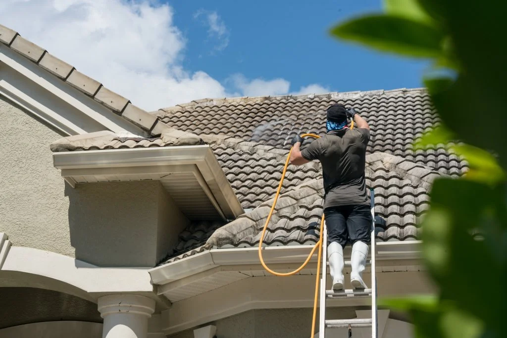 Person on ladder cleaning a residential roof with a hose.
