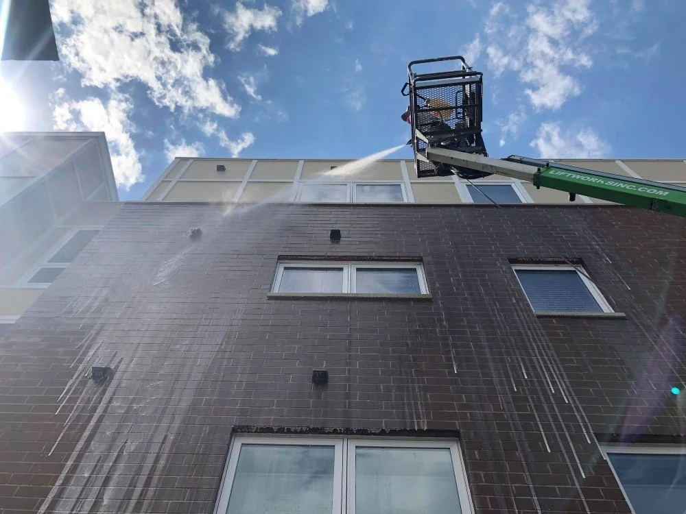 Worker pressure washing building exterior using a boom lift under blue sky.