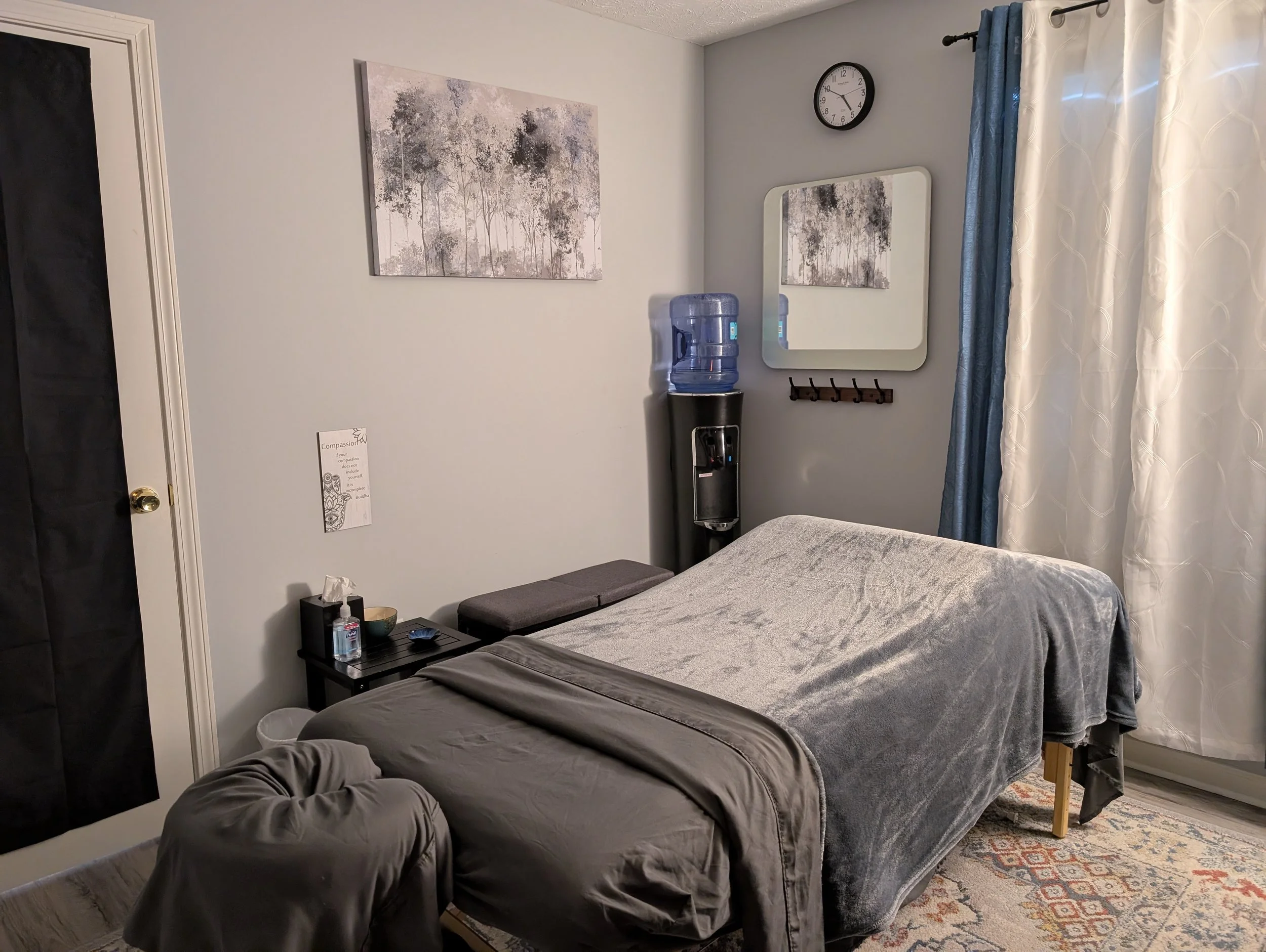 A massage table with a gray blanket, a water cooler, a wall clock, paintings of trees, a mirror, curtains, and a small table with bottle of hand sanitizer and tissues.