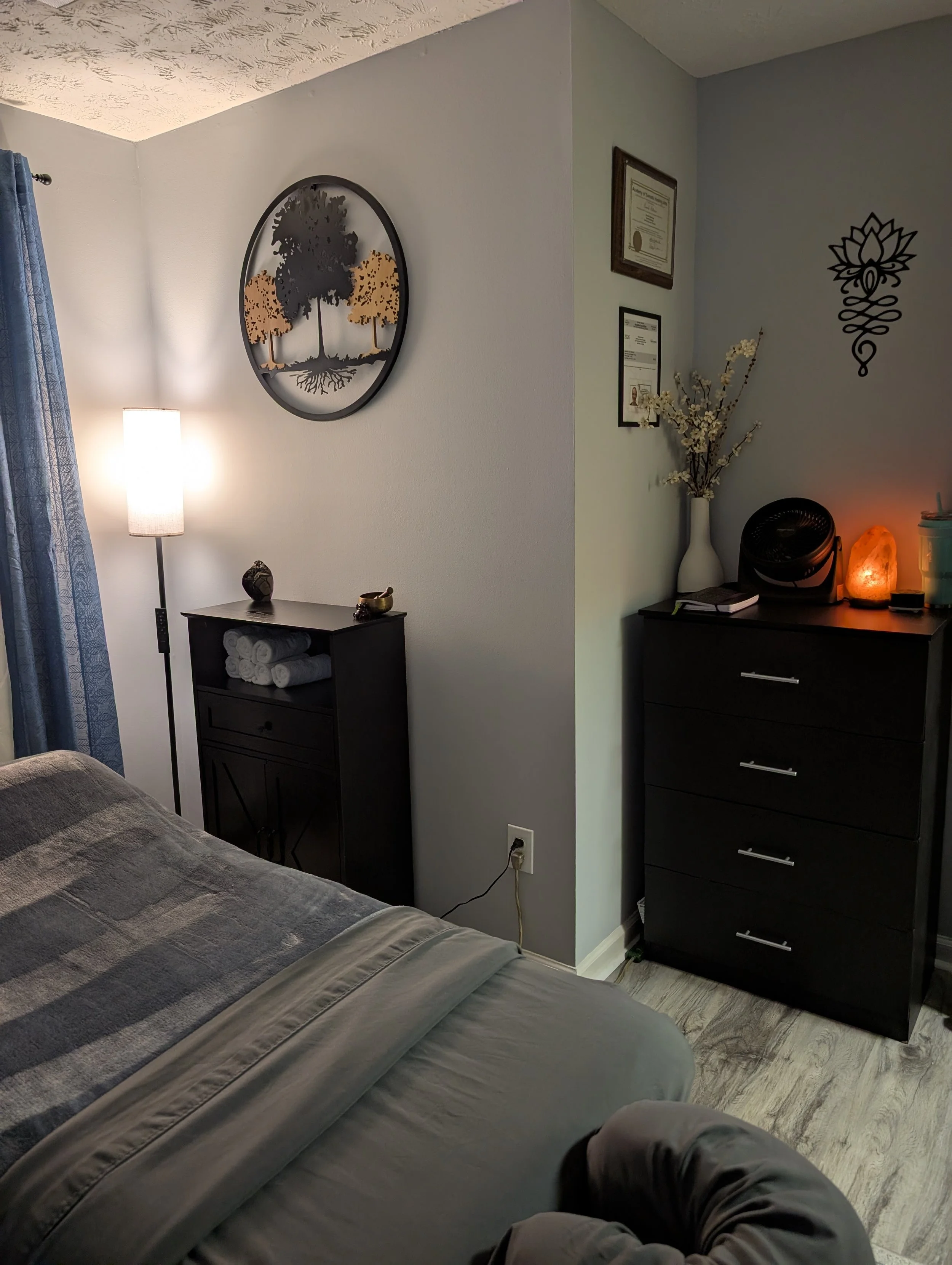 Interior of a massage room showing part of a massage table, a black dresser with rolled towels, a small table, a lamp, wall decorations, framed certificates, a tall vase with white flowers, salt lamp, and a black fan.