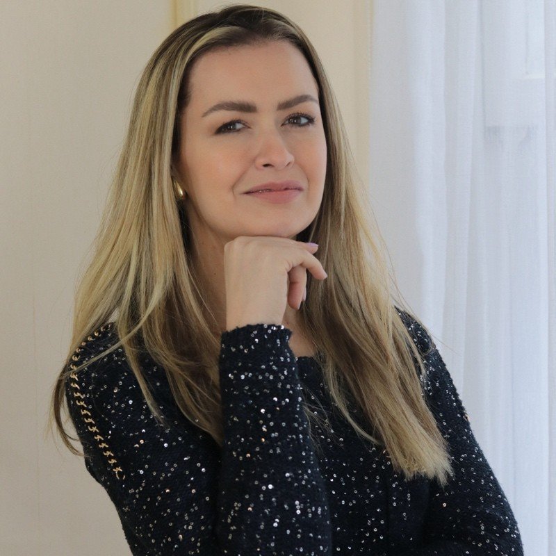 Close-up of a smiling woman with long dark hair, wearing a blazer, with a white brick wall background.