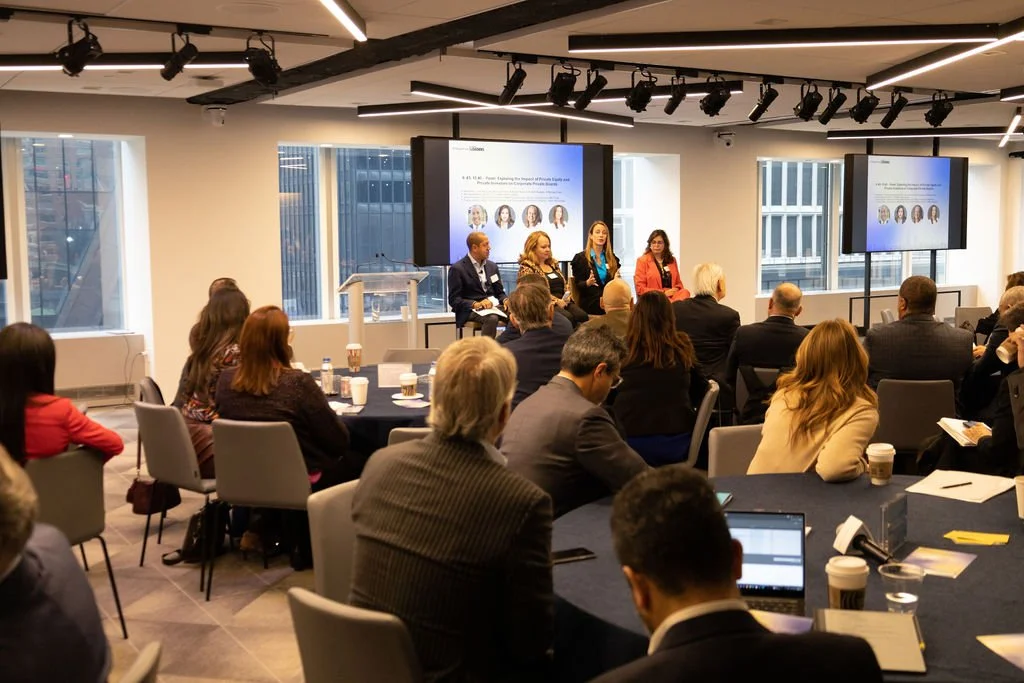 A conference room with a panel of four speakers at the front, a screen displaying a presentation, and an audience seated at round tables, in a high-rise office building with large windows showing city skyscrapers.