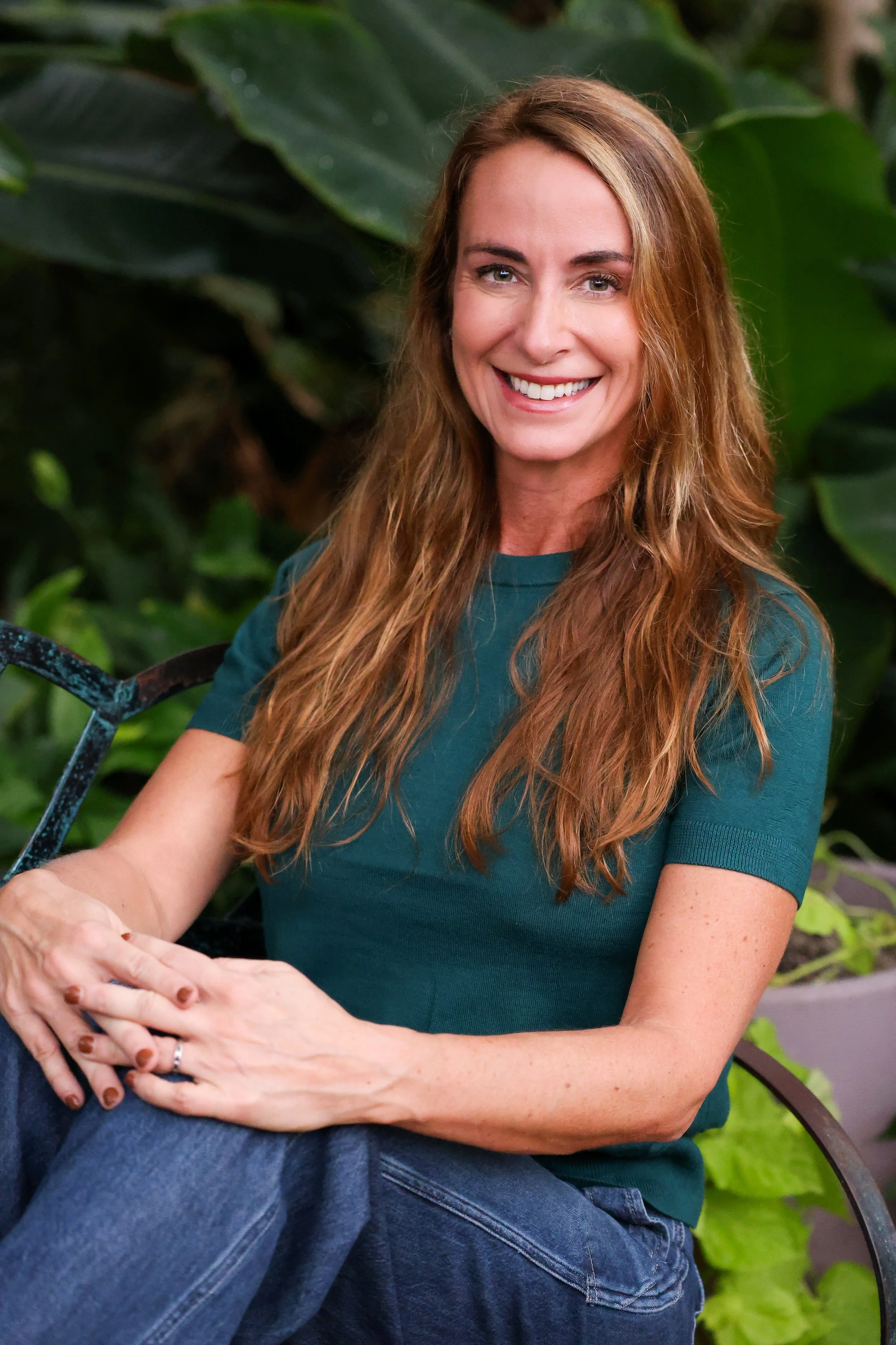 A woman with long, wavy red hair sitting outdoors among green foliage, smiling at the camera, wearing a teal short-sleeve shirt and blue jeans.