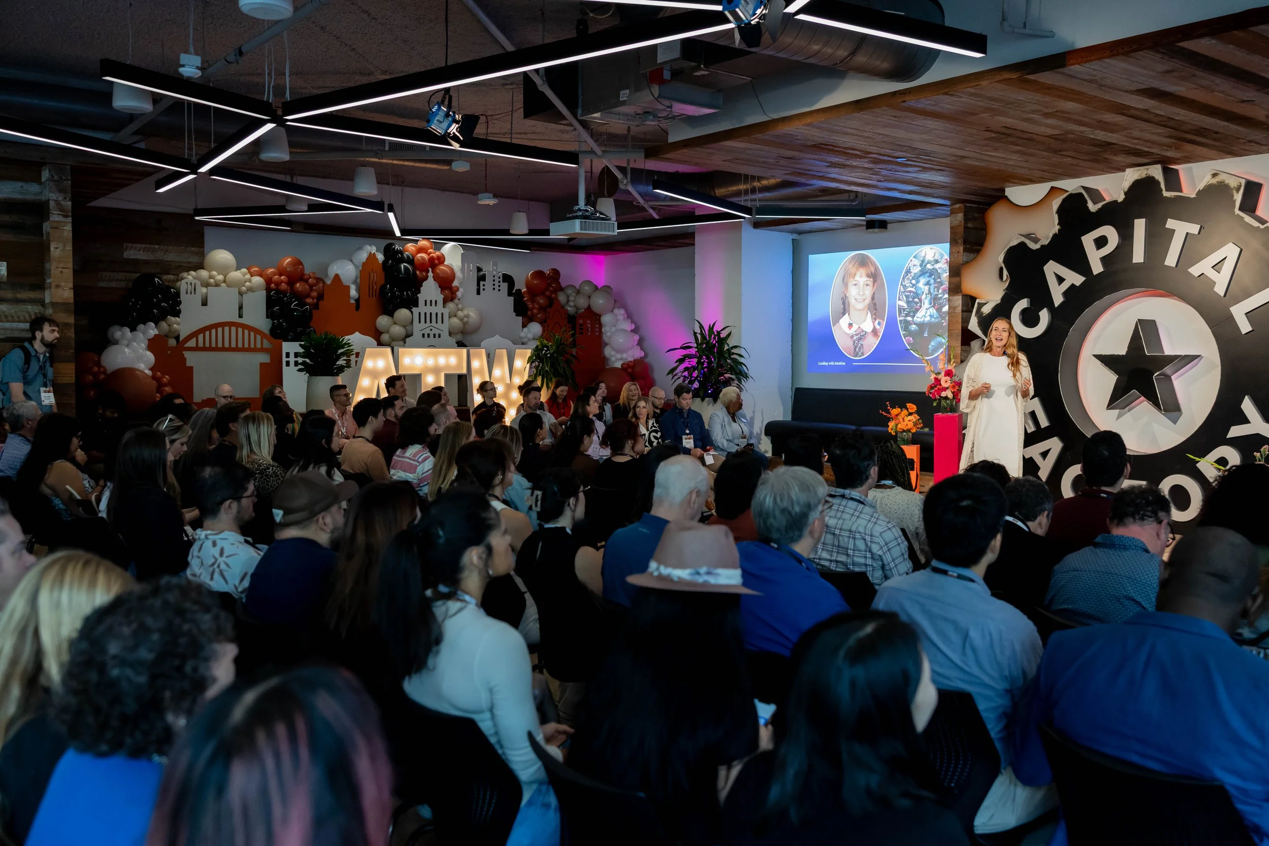 A woman giving a presentation on stage at a Capital Factory event with an audience seated in front. The stage features a large capital factory logo and a screen with a smiling child's photo. Decorative cityscape and balloons are in the background.