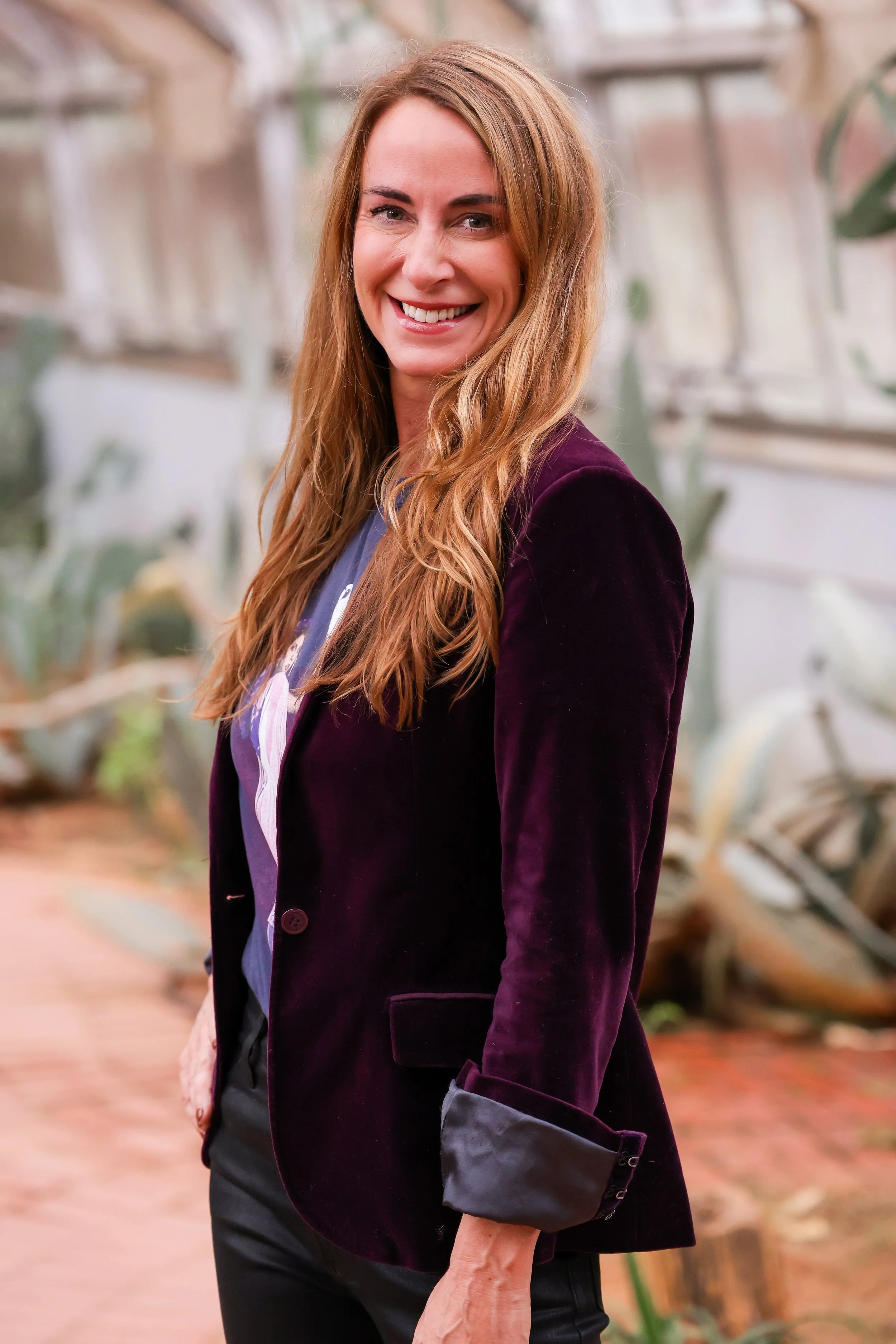A woman with long wavy red hair, smiling and wearing a dark purple blazer over a graphic T-shirt standing indoors with plants and a glass ceiling in the background.