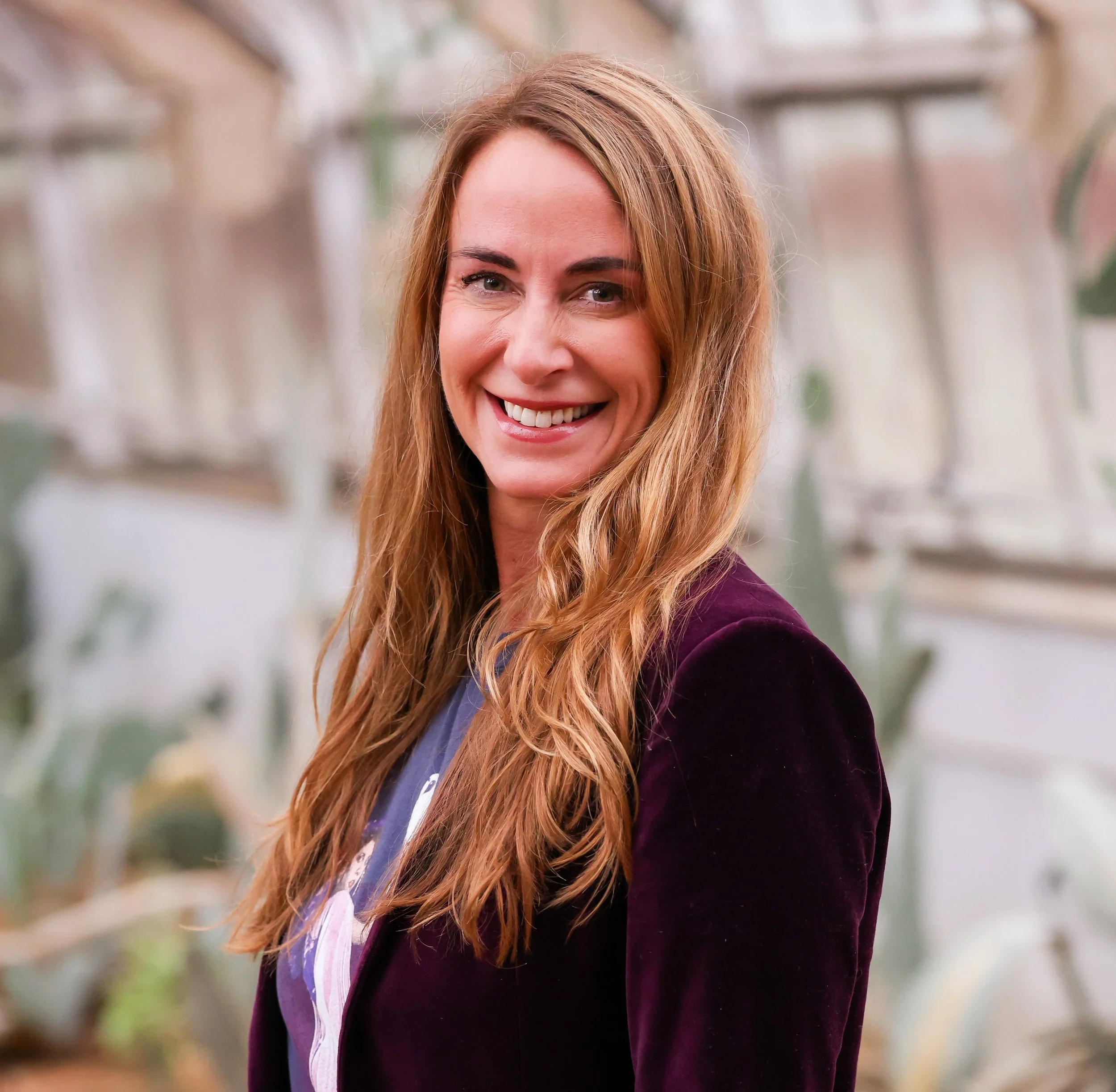 A woman with long, wavy red hair smiling at the camera, wearing a dark purple blazer over a patterned blouse, with blurred greenhouse plants in the background.