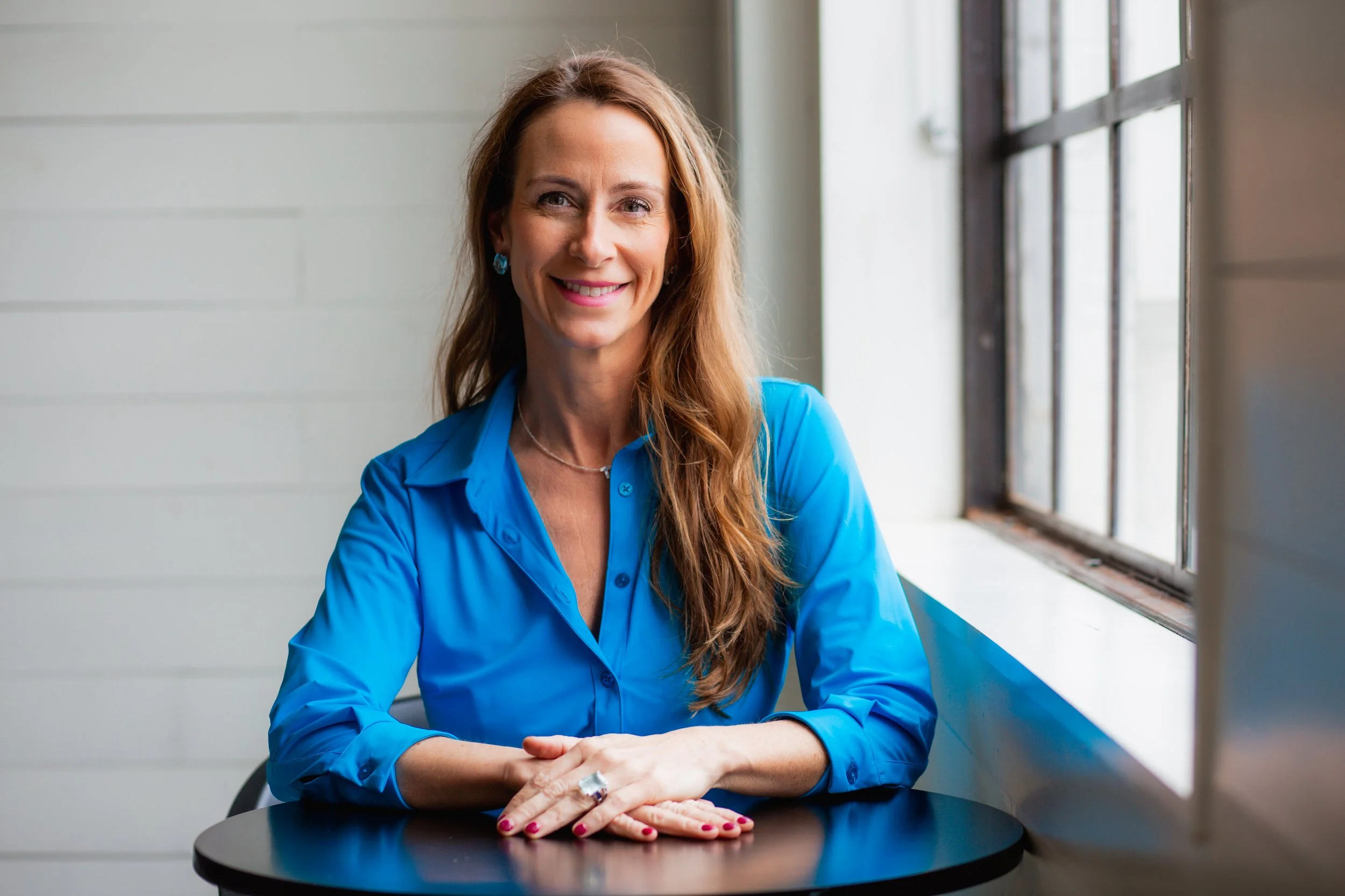 Tracey Abbott seated at a table with hands clasped, wearing a blue blouse, near a window.