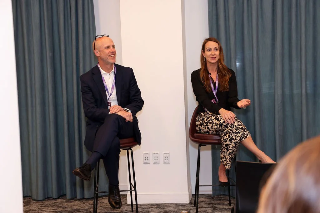 Two people sitting on high chairs in conversation in front of teal curtains during a presentation or panel discussion.