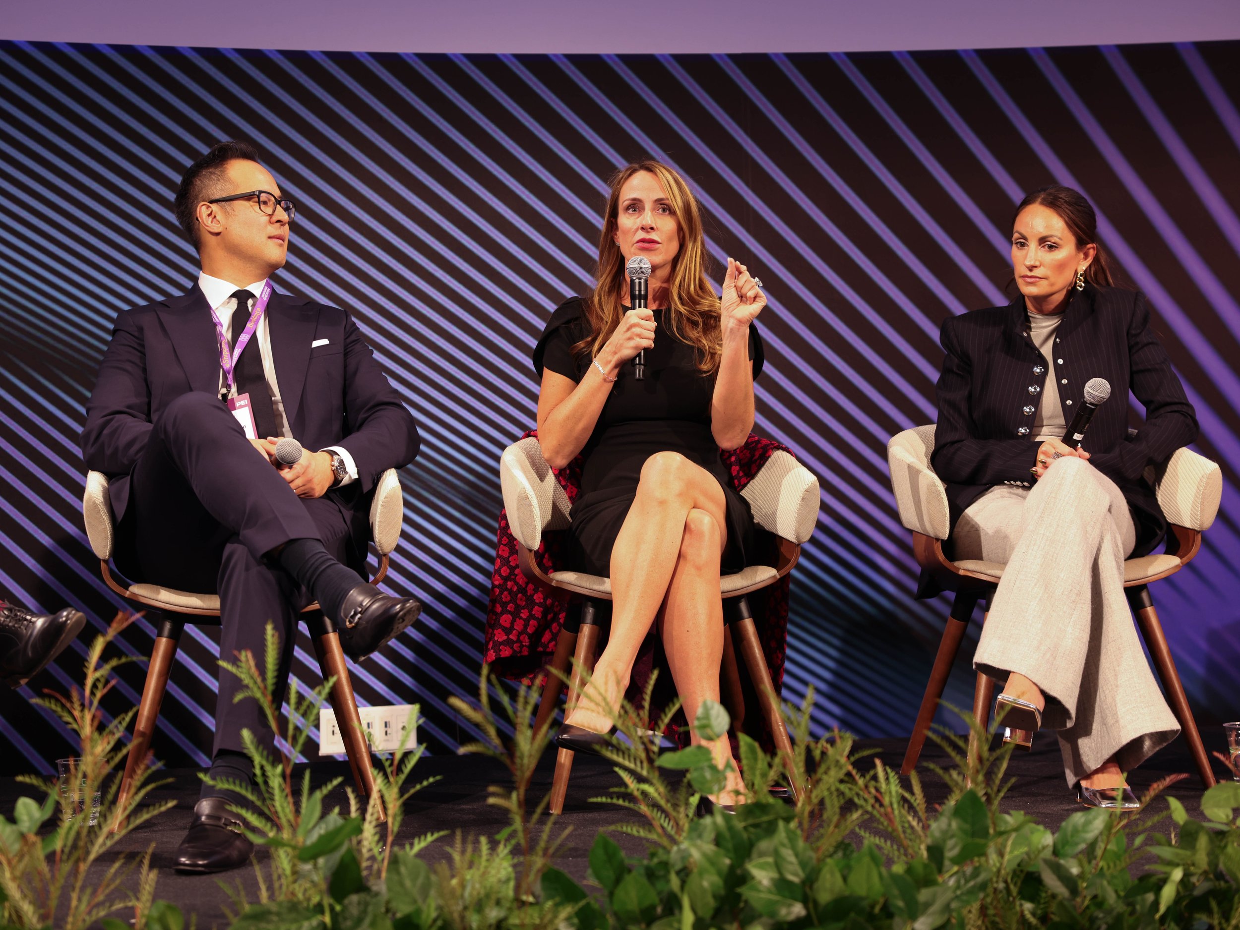Three people seated on a stage during a panel discussion, with a man on the left wearing glasses, a woman in the middle speaking into a microphone, and another woman on the right holding a microphone. The background features diagonal purple and black lines, and there are plants at the front of the stage.