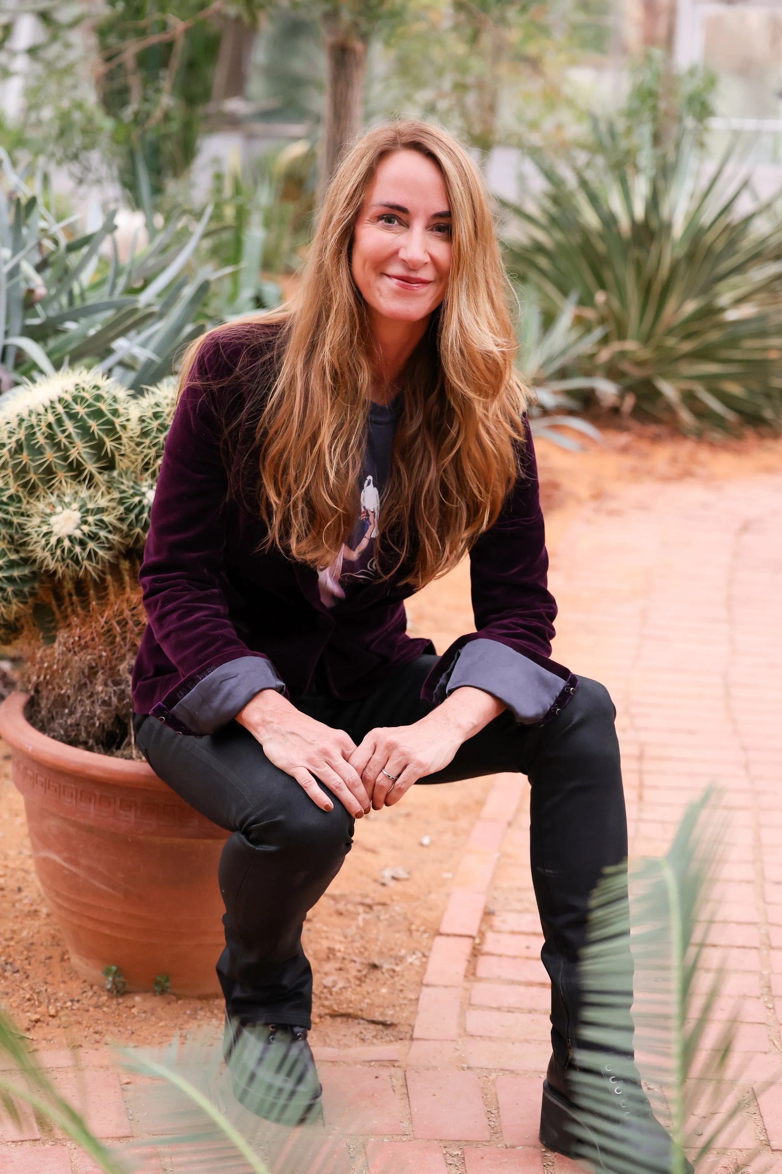 A woman with long, wavy, reddish-brown hair sitting on a brick path beside a large pot of cactus plants in a greenhouse or botanical garden.
