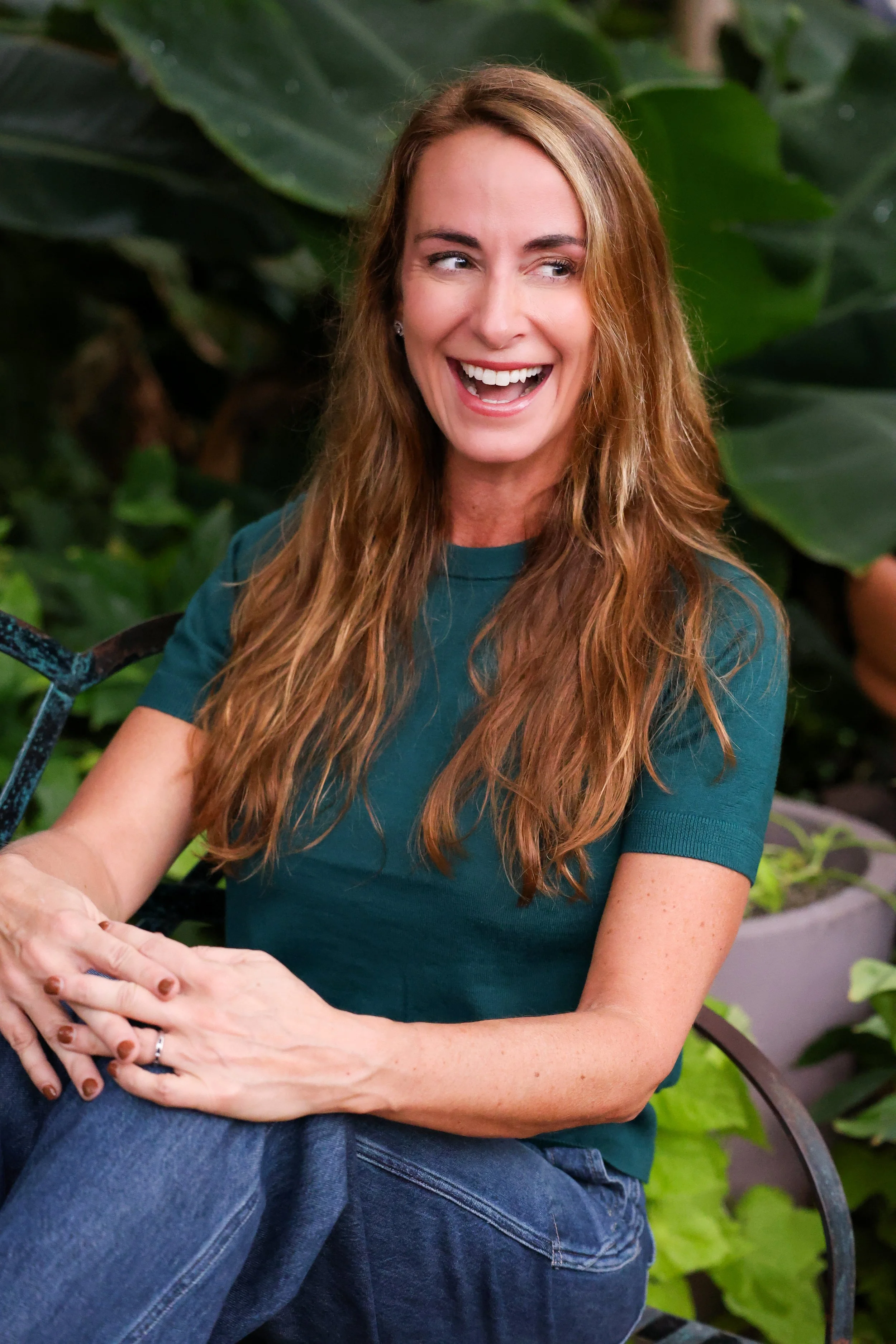 A woman with long, wavy red hair sitting outdoors on a black metal chair, smiling and looking to her right, with large green leafy plants in the background.