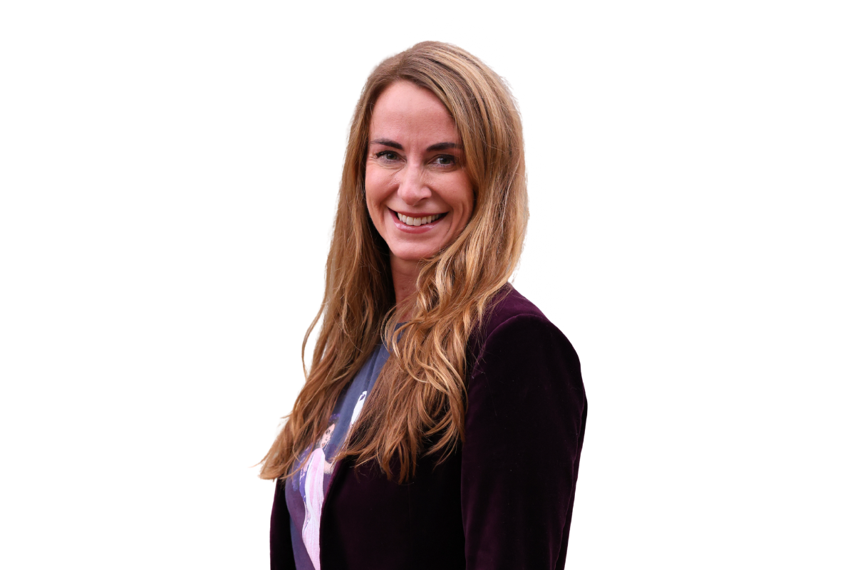 Smiling woman with long, wavy, reddish-brown hair wearing a black blazer and a graphic t-shirt against a white background.