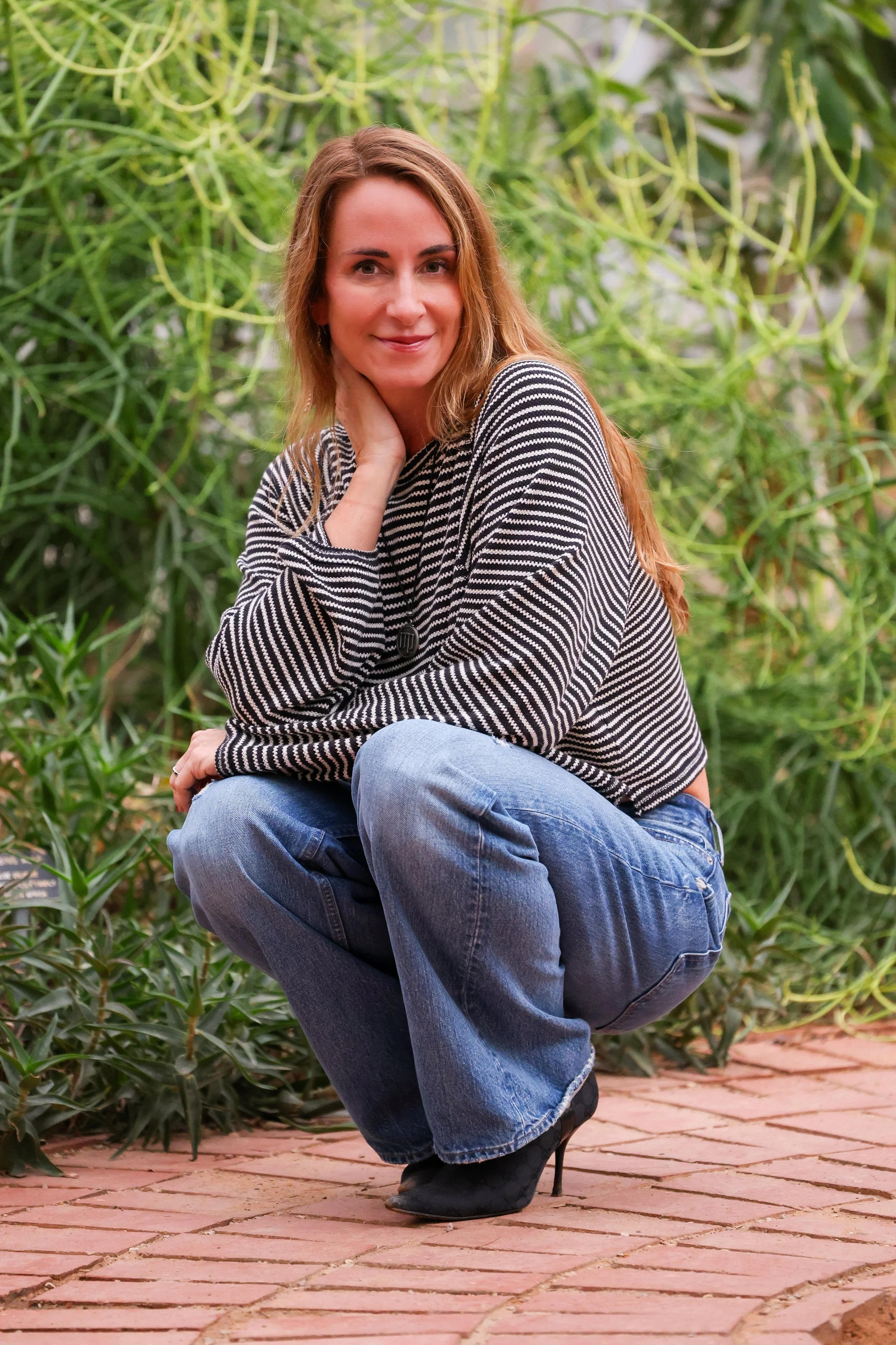 A woman squatting on a brick pathway outdoors, surrounded by greenery, with a slight smile, wearing a black and white striped sweater, blue jeans, and black high heels.