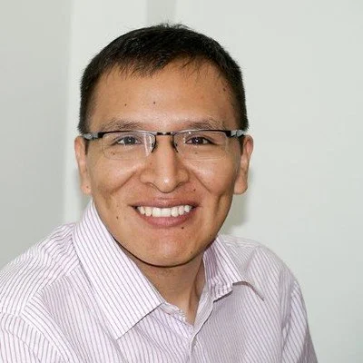 Headshot of a man with light brown hair, blue eyes, wearing a navy blazer and light blue shirt, smiling against a gray background.