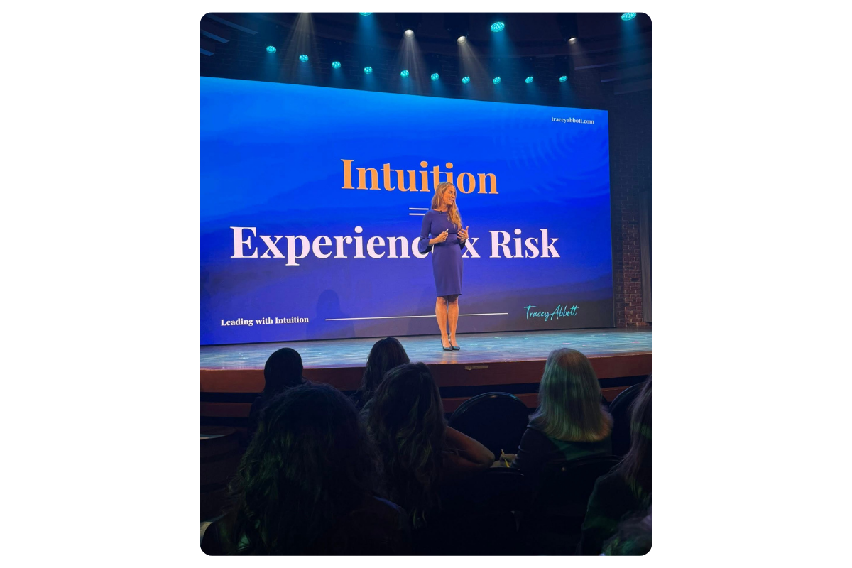 A woman giving a presentation on a stage with a large screen behind her displaying the words "Intuition = Experience x Risk". The audience is seated in front of the stage, watching her.
